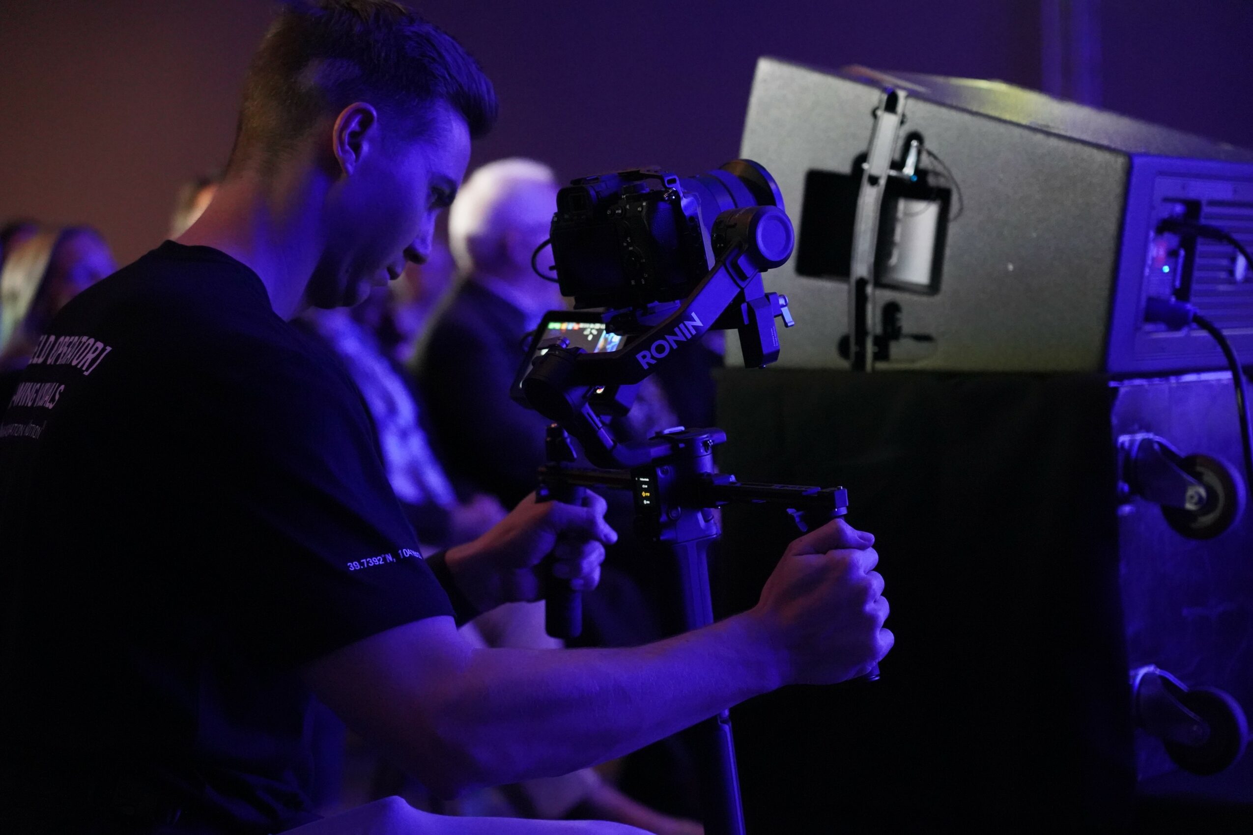 Person operating a camera on a tripod in a dark room with equipment nearby.