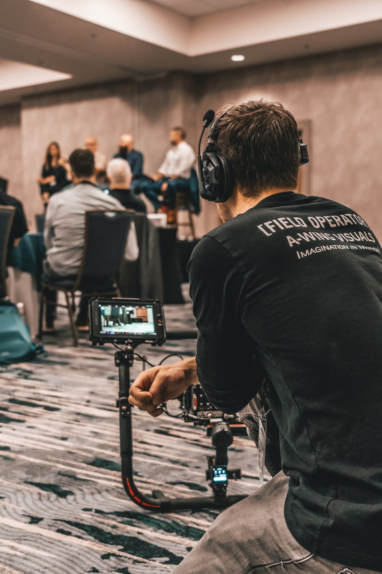 Person operating camera equipment in a conference room with audience and speaker in background.