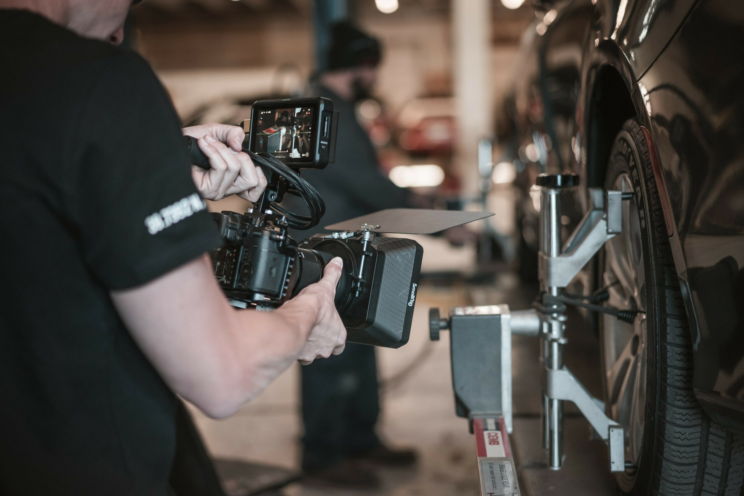 Person holding a professional camera, taking photos of a car in a garage or workshop setting.