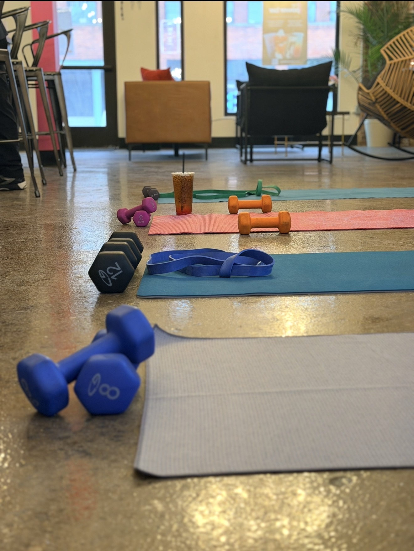 Yoga mats, dumbbells, resistance bands, and water bottles arranged on a wooden floor in a room with large windows and seating area.