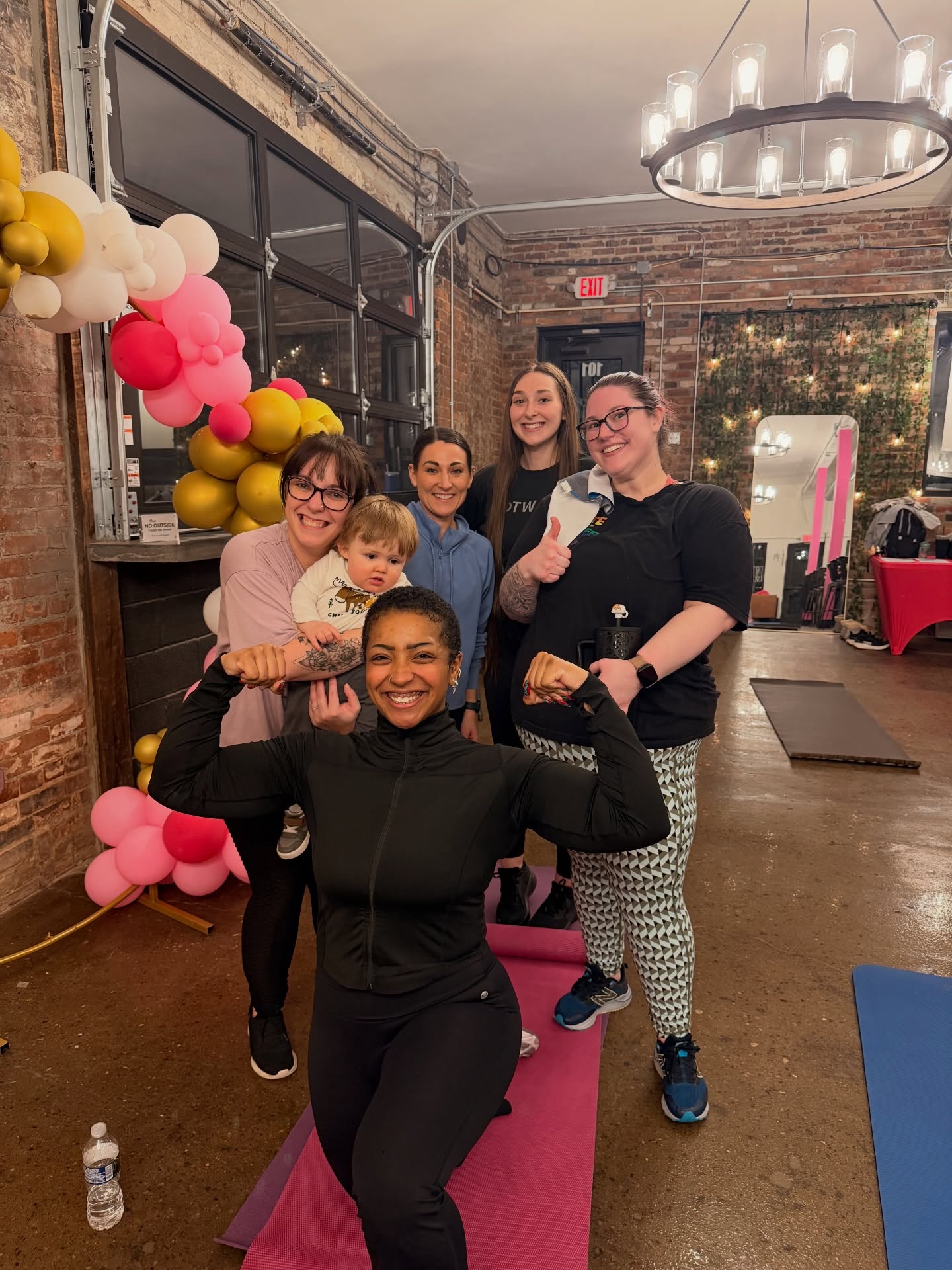 Group of six women and children smiling indoors, some making gestures, with balloons and brick wall in background.