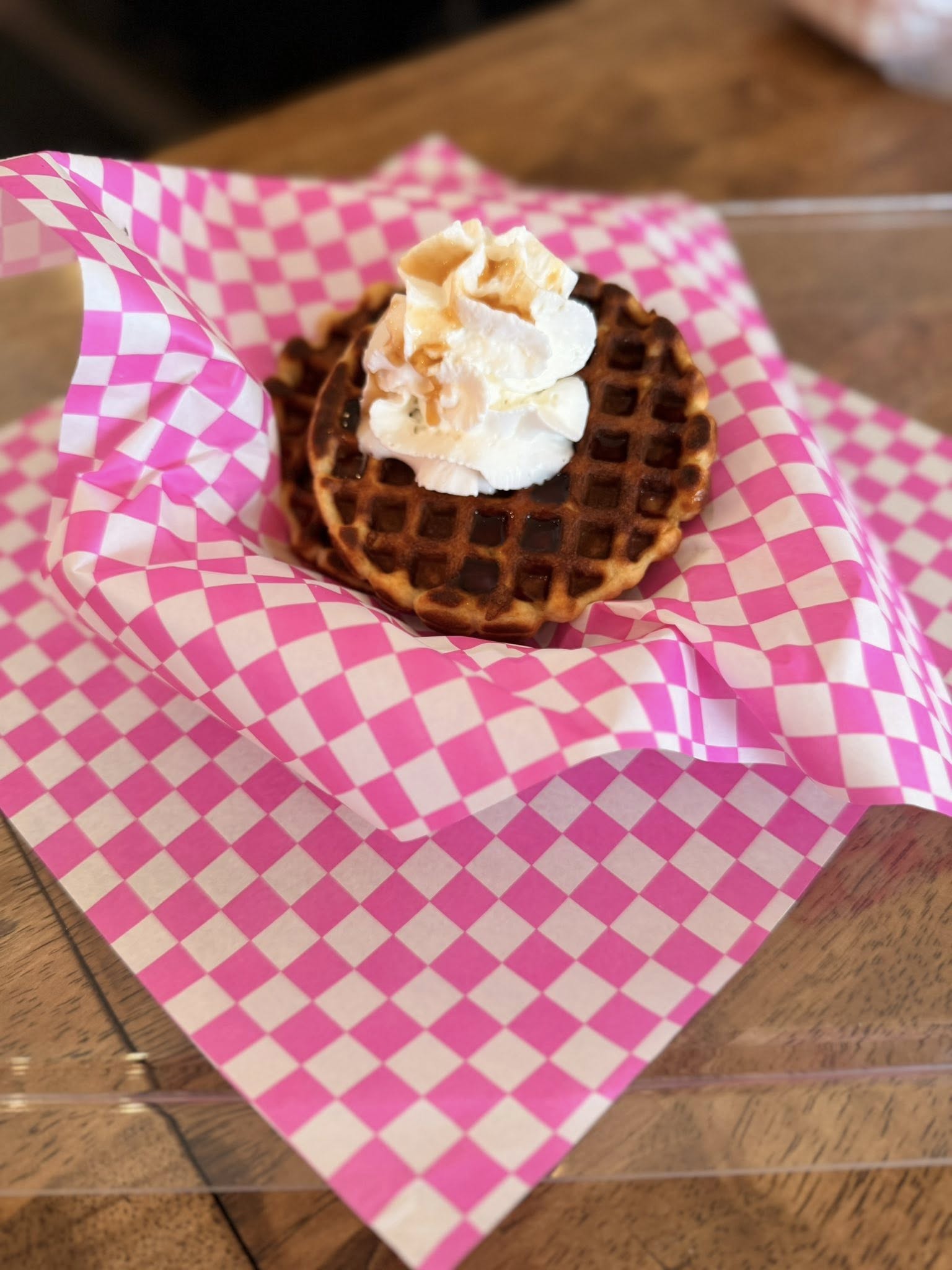 Waffle topped with whipped cream on pink checkered paper, on wooden table.
