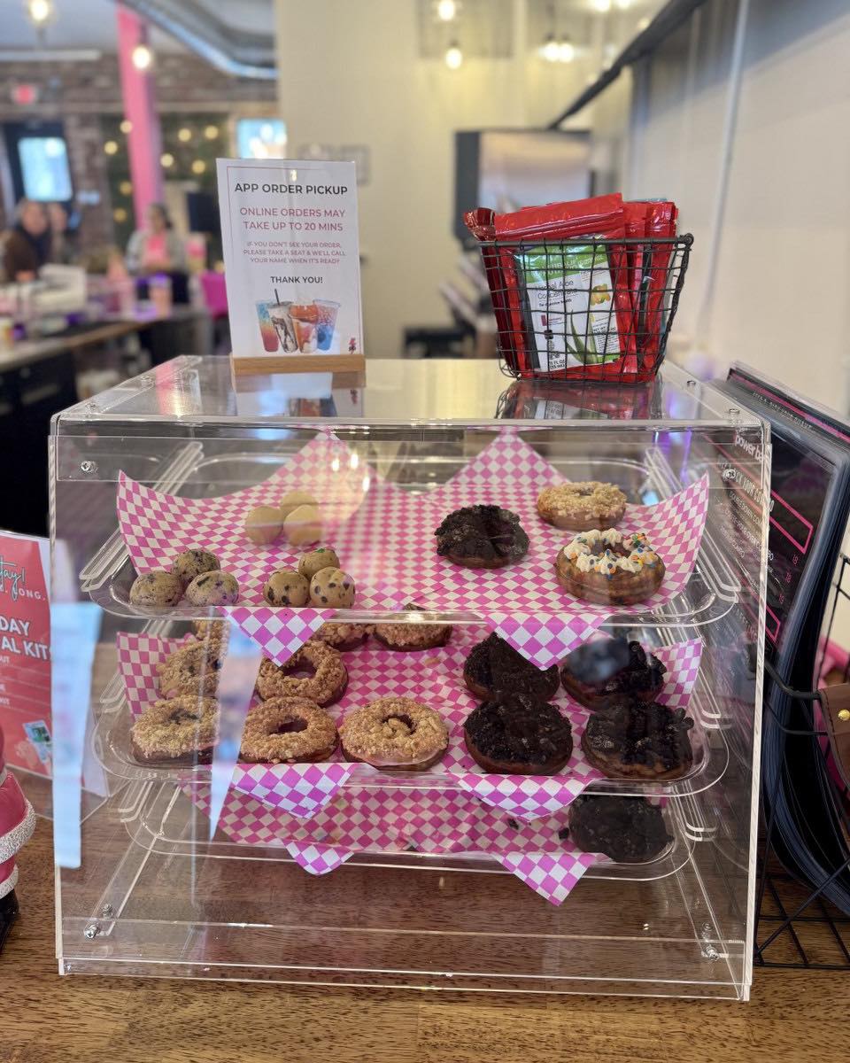 Clear display case with assorted cookies on pink checkered paper, shopping basket and sign in background.