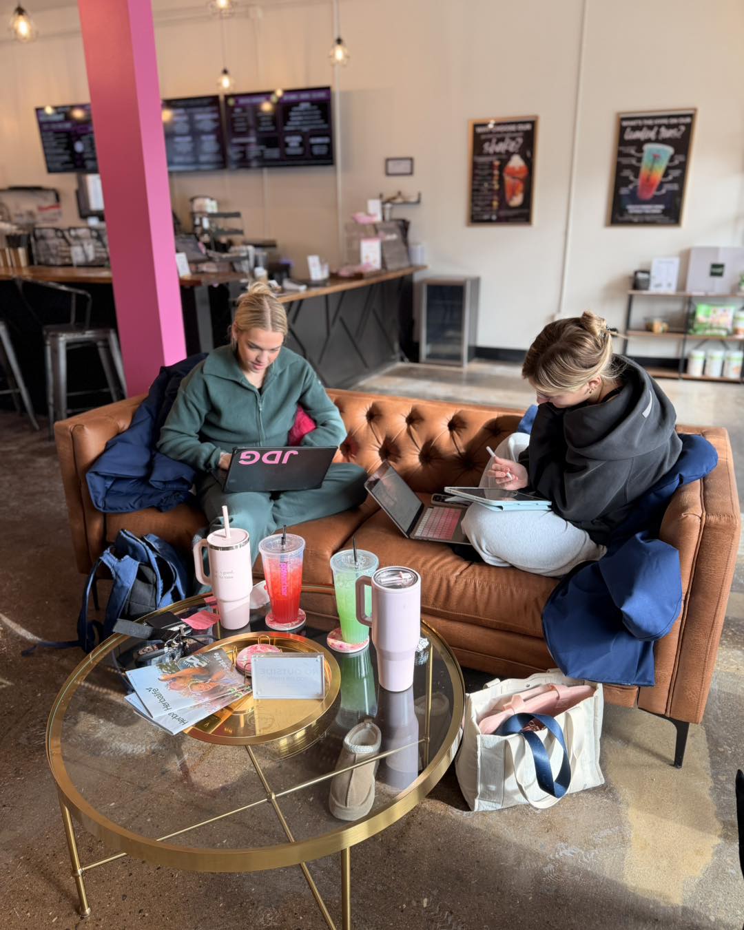 Two women sit on a brown sofa in a cafe, using laptops and surrounded by drinks and bags on a glass table.