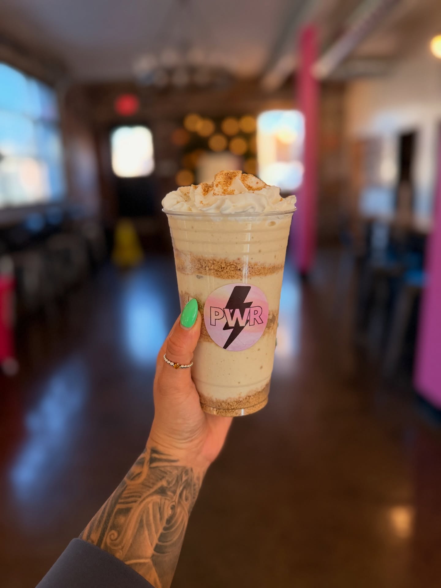 Hand holding a blended coffee drink topped with whipped cream inside a cafe with blurred background.
