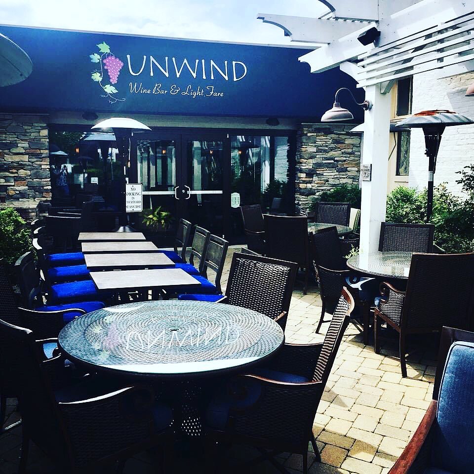 Outdoor seating area with tables and chairs outside a restaurant named UNWIND, with umbrellas and a stone wall.