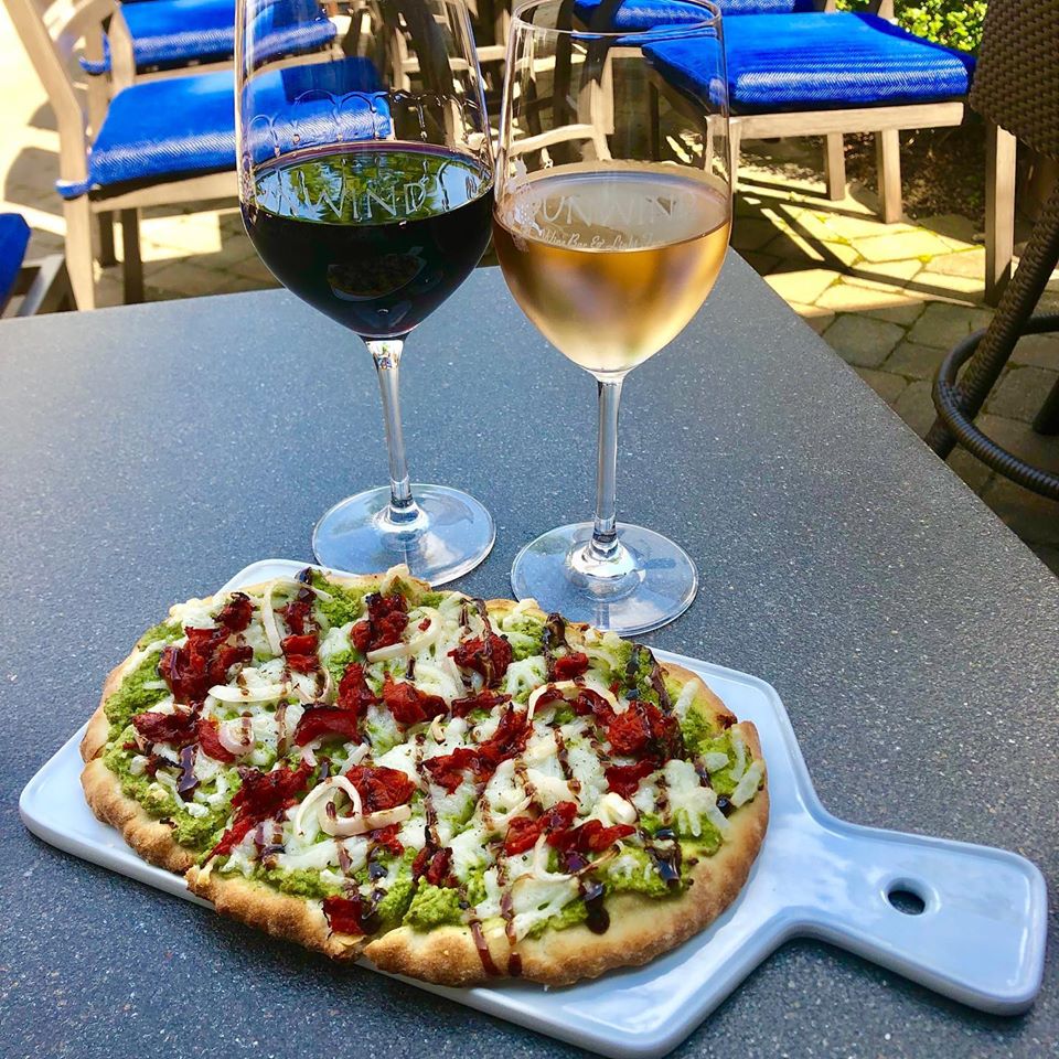 Pizza with green topping, cheese, and red sauce on a white serving board, two glasses of wine on a dark table outdoors.