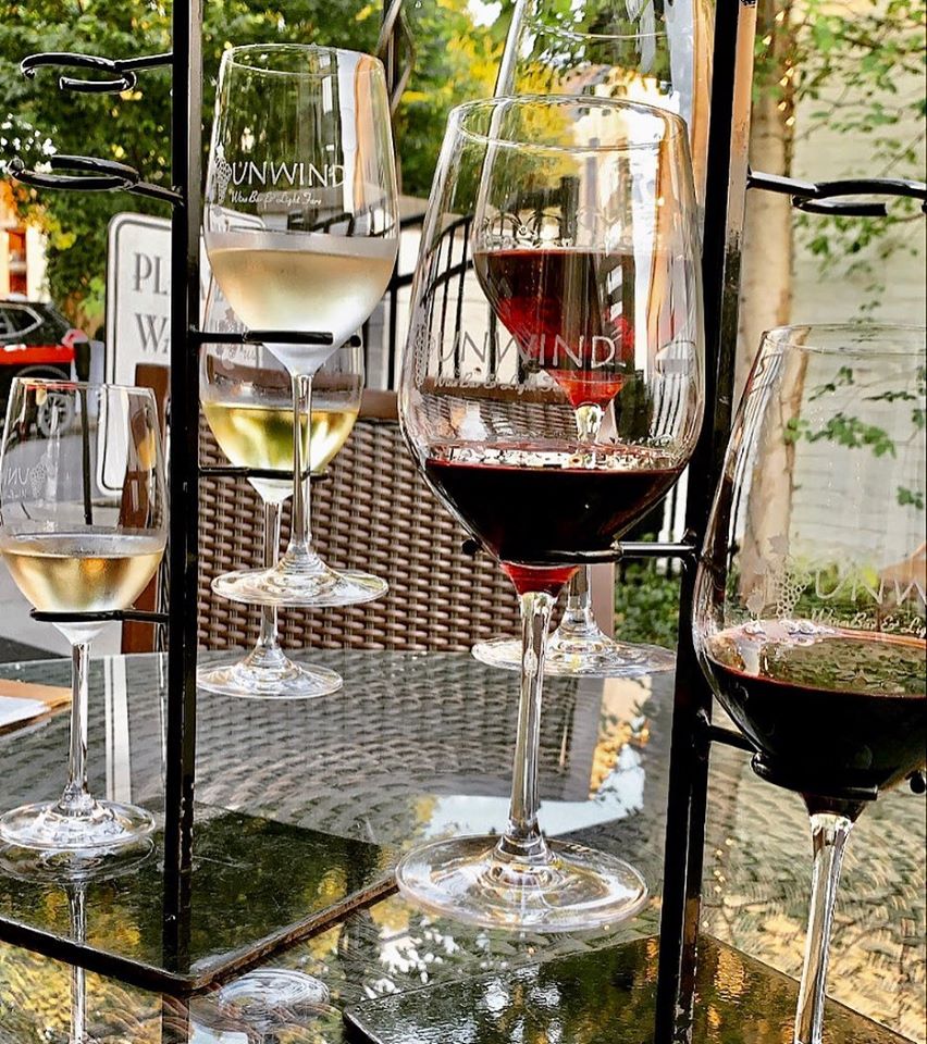 Four wine glasses with white, red, and rosé wines on a glass table outdoors, with trees and a fence in background.