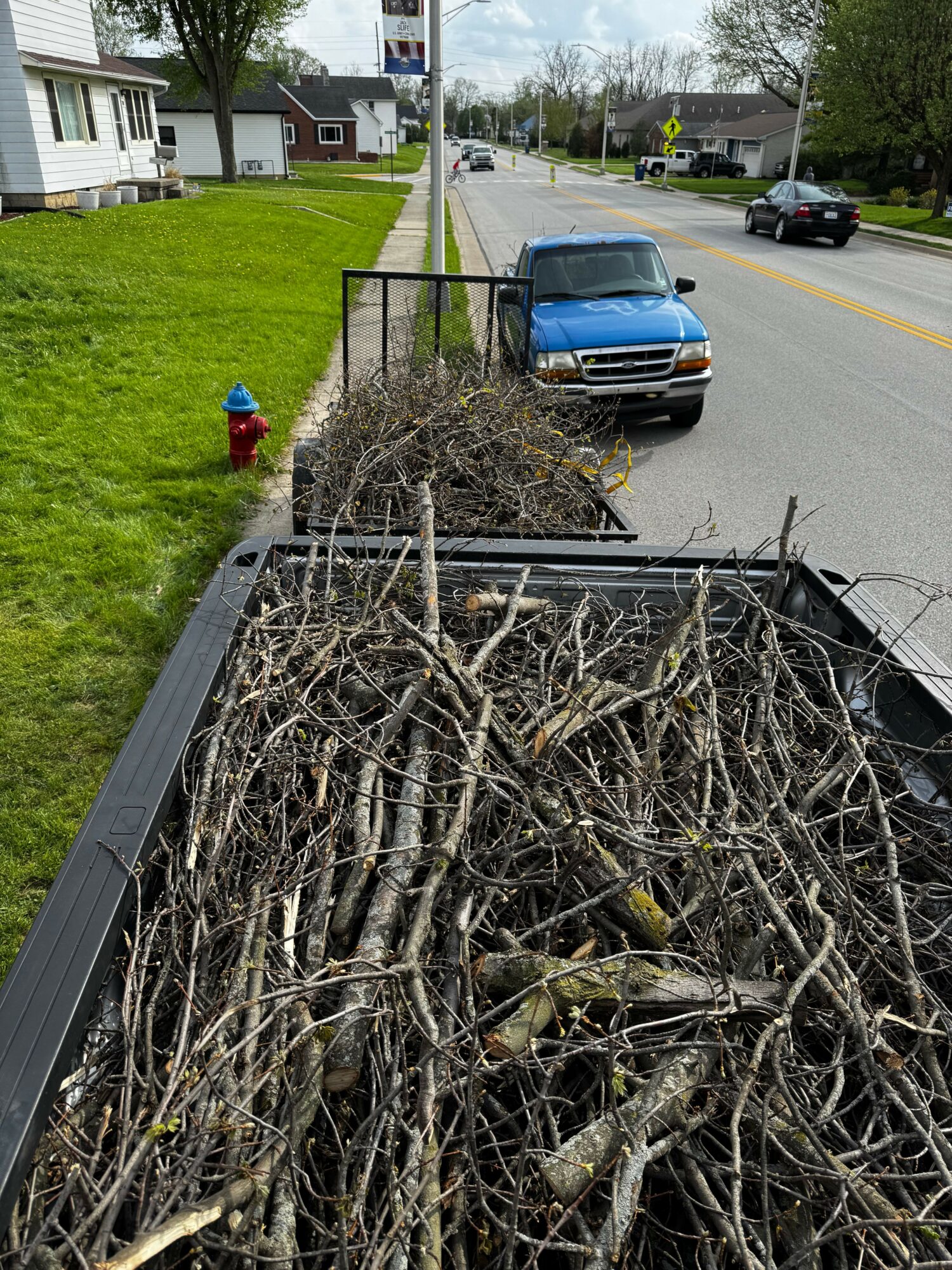 View of a street with a parked blue car, a pile of branches in a truck bed, and houses with green lawns.
