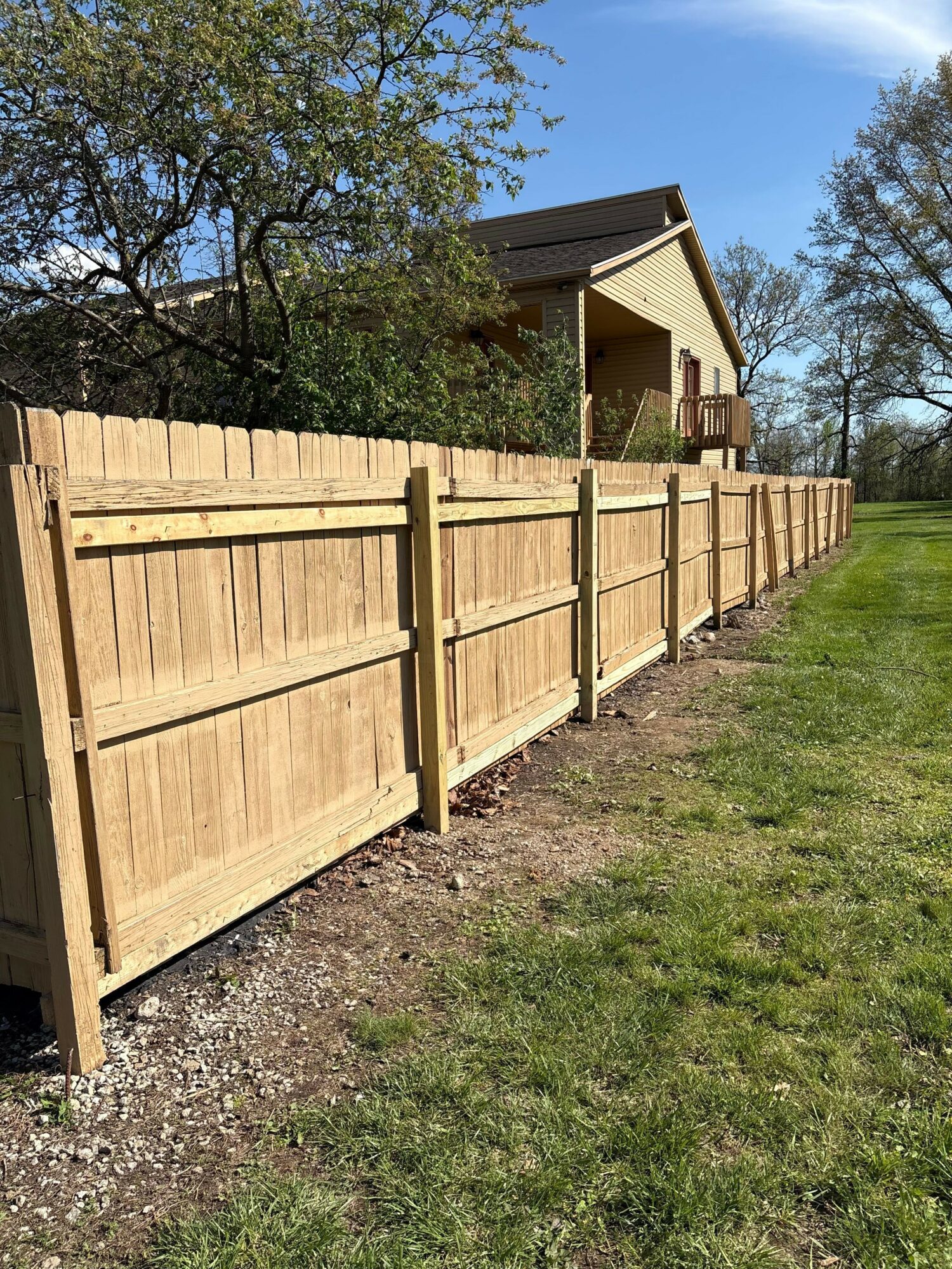 Wooden fence runs along a grassy yard with a house and trees in the background under a blue sky.