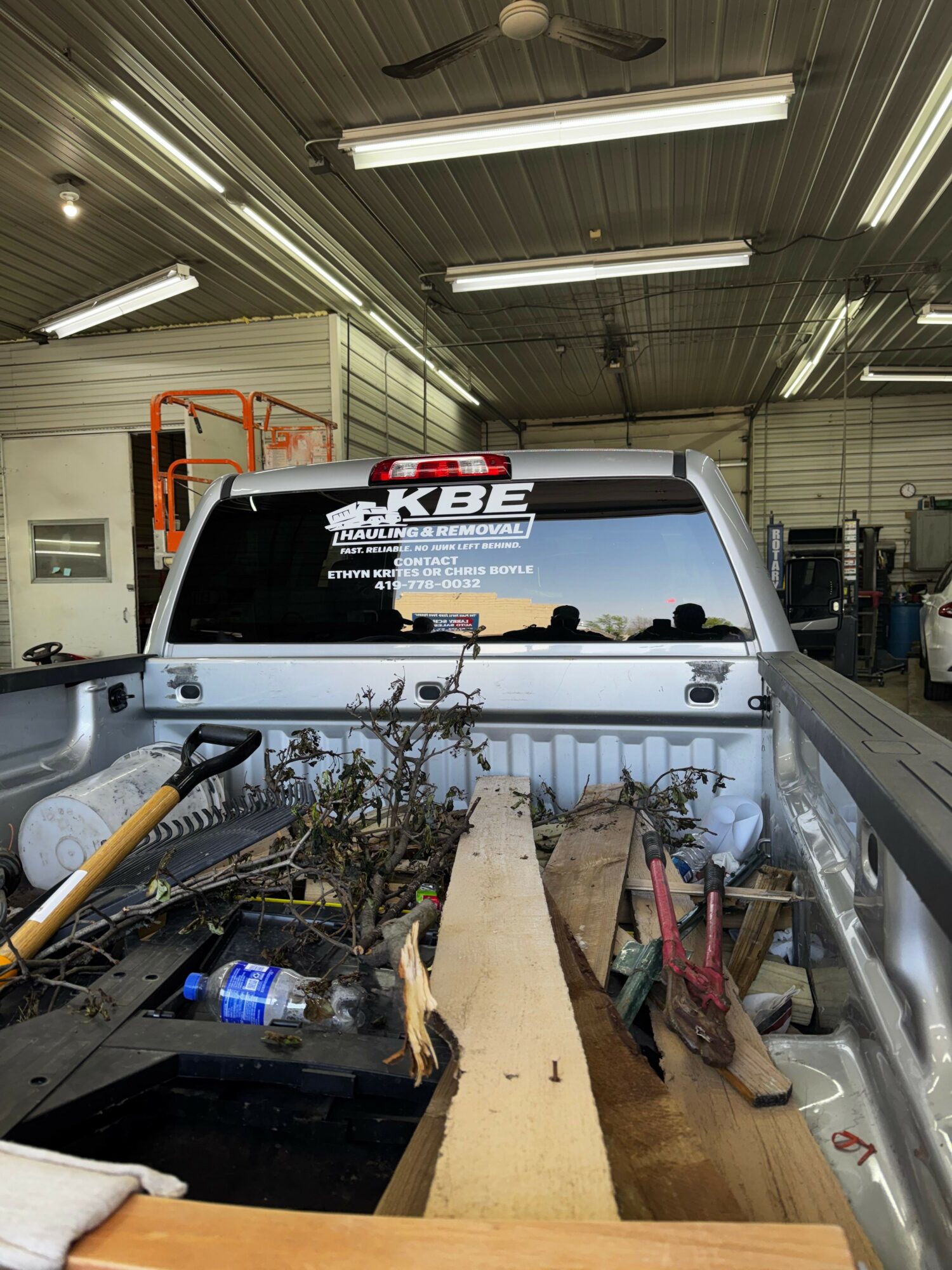 View of a pickup truck bed with tools, wood, and plants inside, inside a workshop with fluorescent lights and metal ceiling.