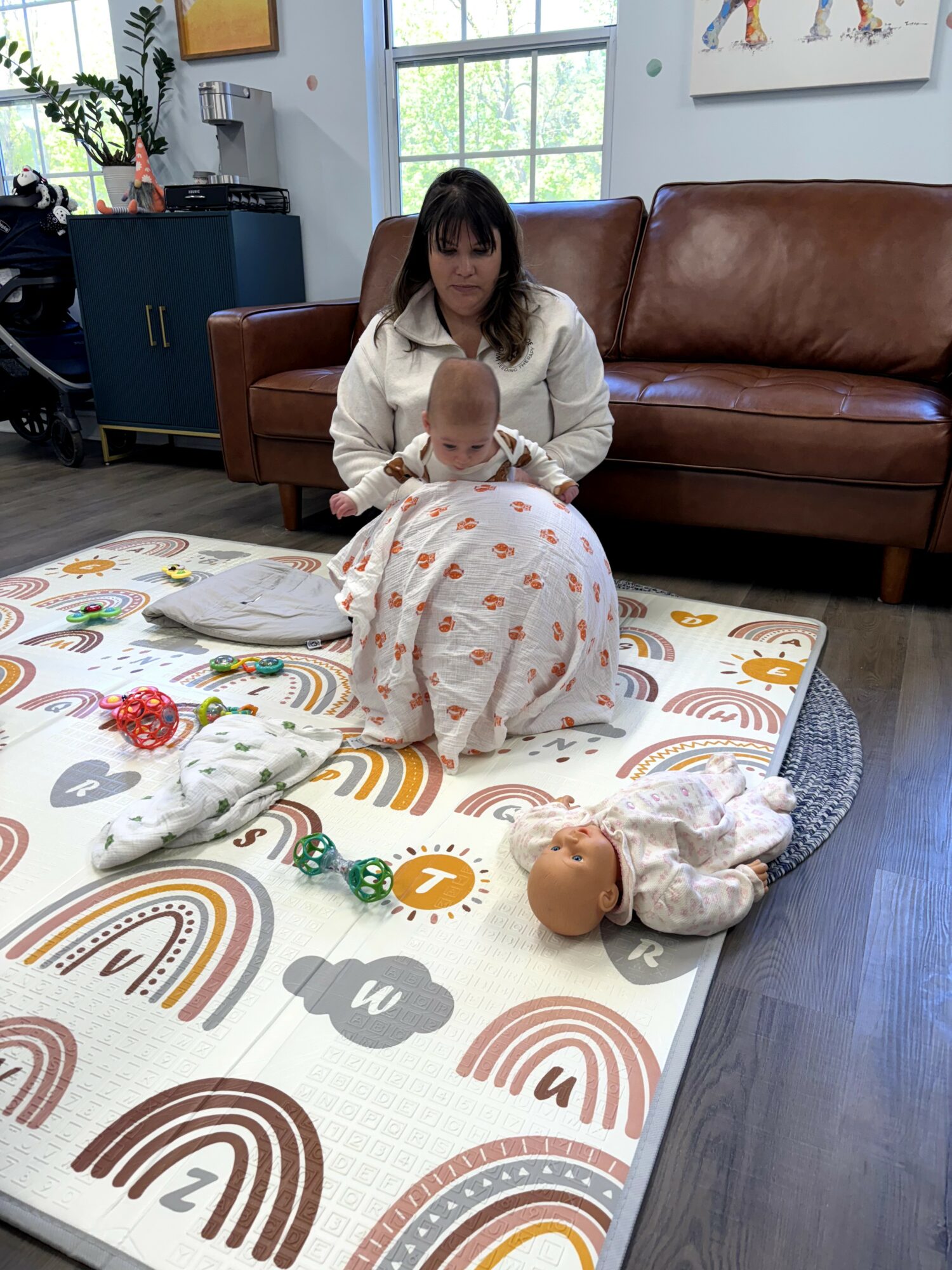 Woman sitting on a couch with a baby on her lap, toys and dolls on a colorful mat, in a room with large windows.
