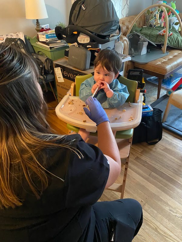 Child sitting in high chair drinking from a cup, caregiver wearing gloves assisting, room with furniture and toys in background.