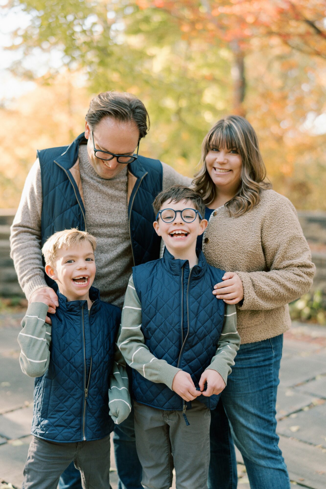 Family of four outdoors in autumn, smiling, with trees in background, two children and two adults standing close together.