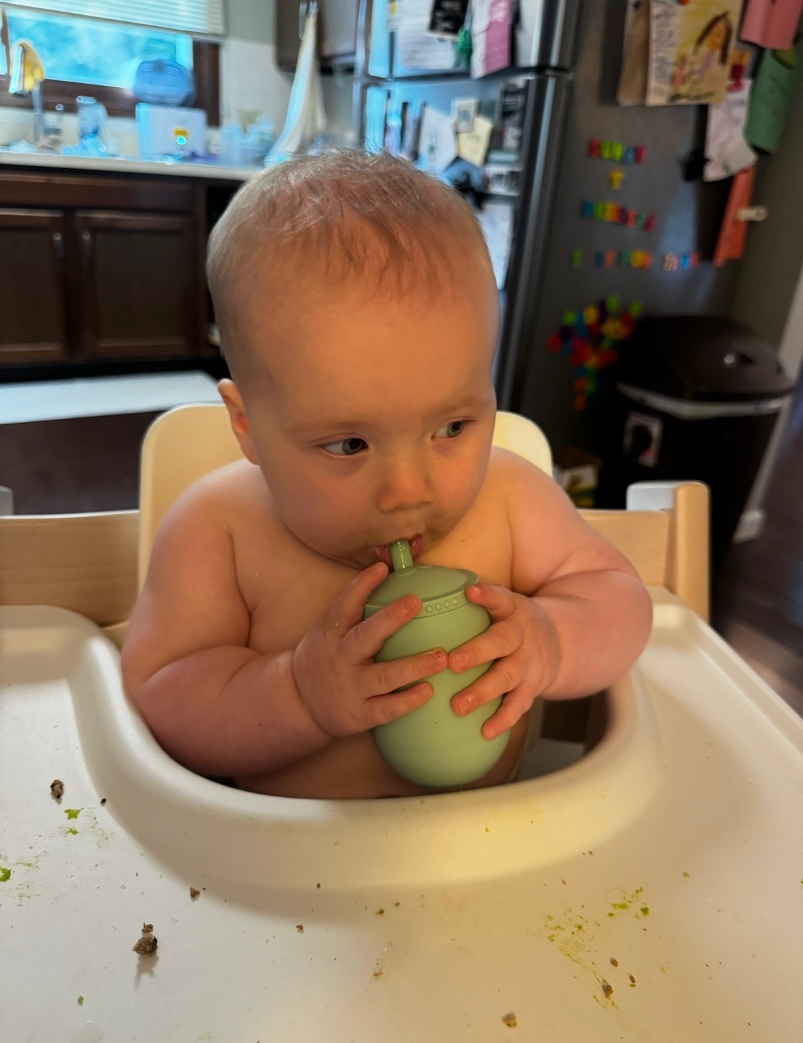 Baby sitting in high chair holding a green cup, kitchen background with refrigerator and cabinets.