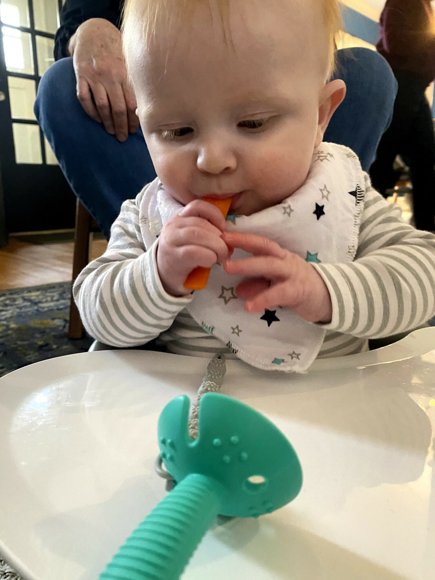 Young child with blond hair and striped shirt holding a small object, sitting at a table with a pacifier in front.