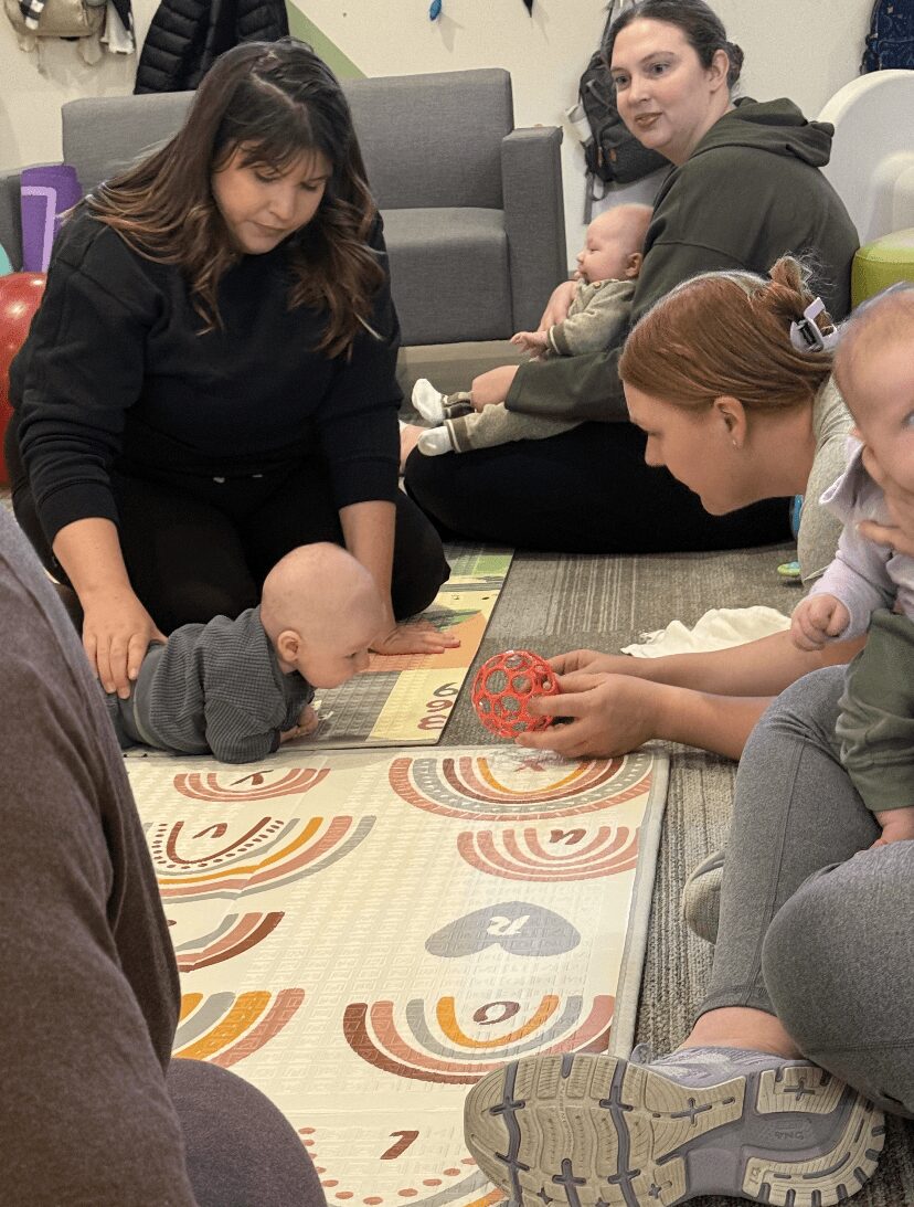 Children and adults playing a board game on the floor indoors, with some sitting on a couch in the background.