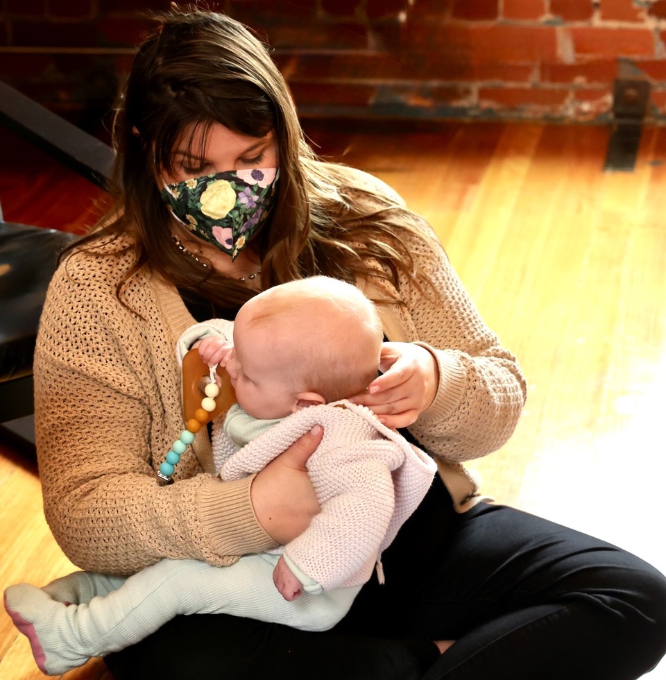 Woman wearing a floral face mask holding a baby with a pacifier, sitting on a wooden floor with a brick wall background.