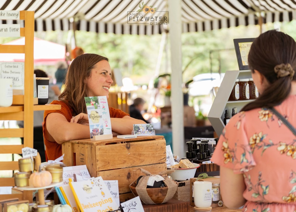 Two women at a market stall, one smiling, with various products and a striped canopy overhead.