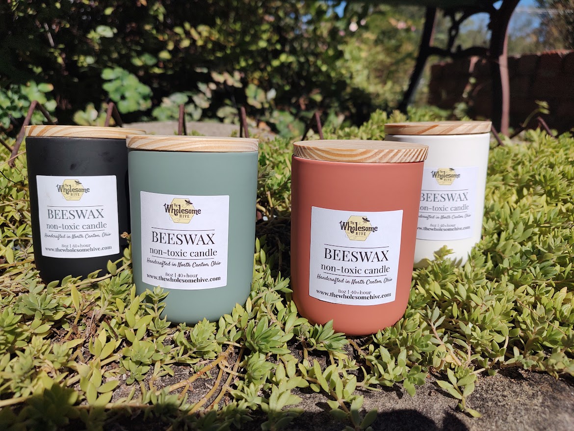 Three candles in different colored containers placed on green plants outdoors, with trees and a brick wall in background.