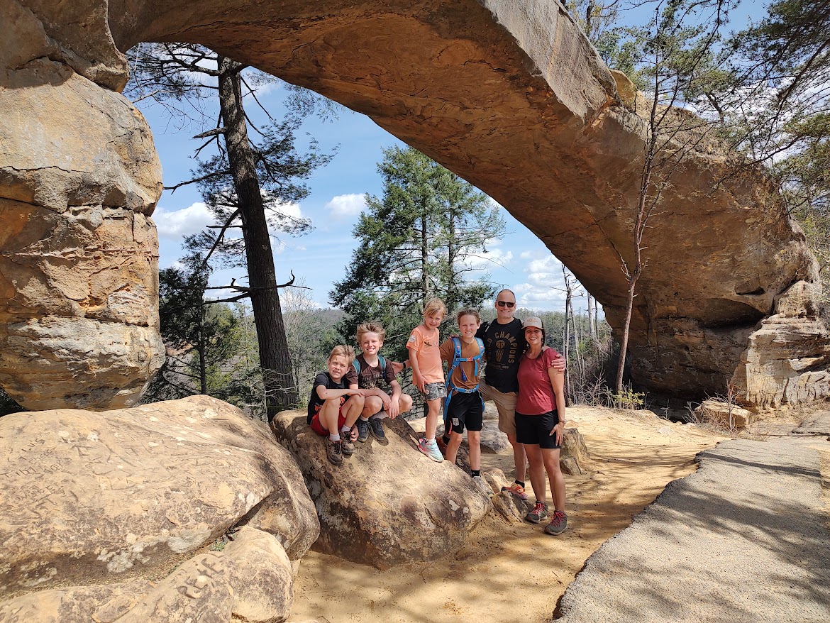 Group of six children standing and sitting on rocks under a large natural rock arch in a forested area.