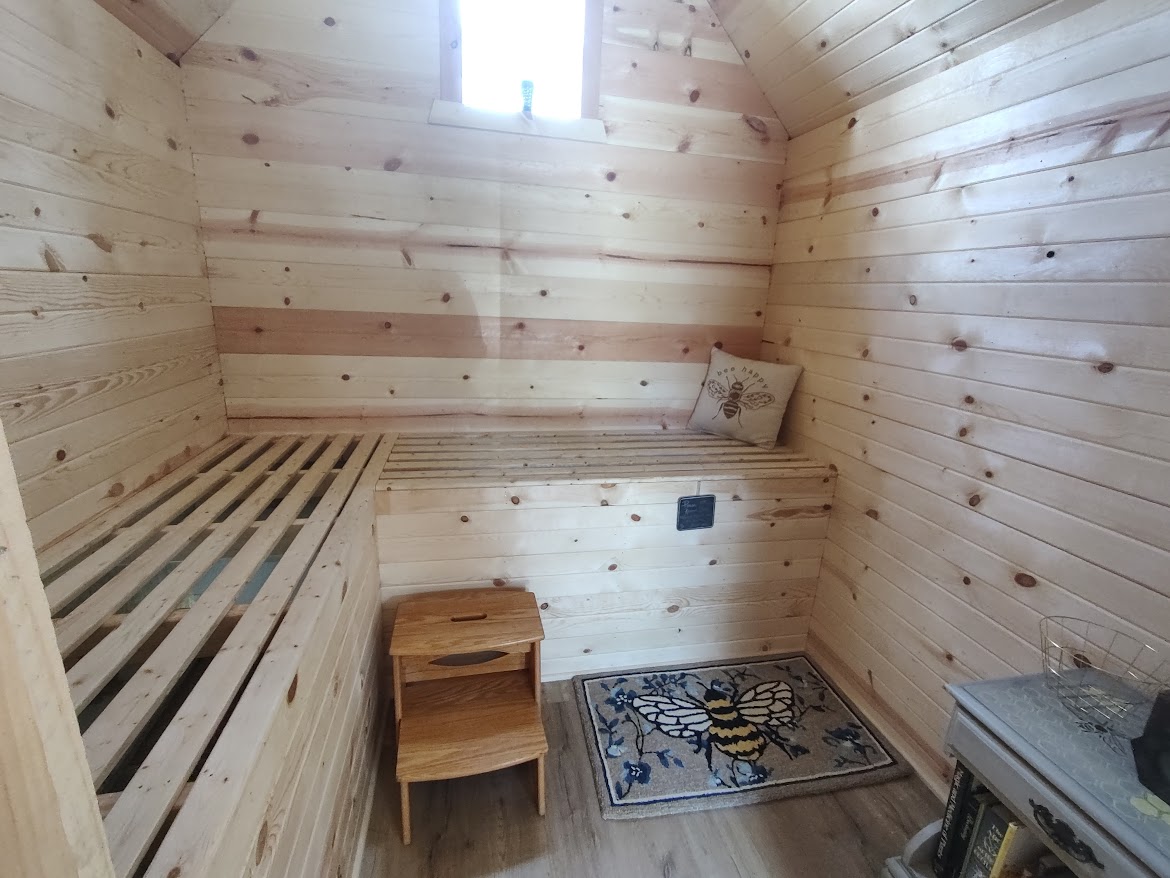 Small wooden room with a window, a bed frame, a small wooden table, and a patterned rug on the floor.