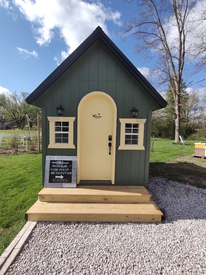 Small green playhouse with cream door and window frames, two black lanterns, wooden steps, and gravel path in front.