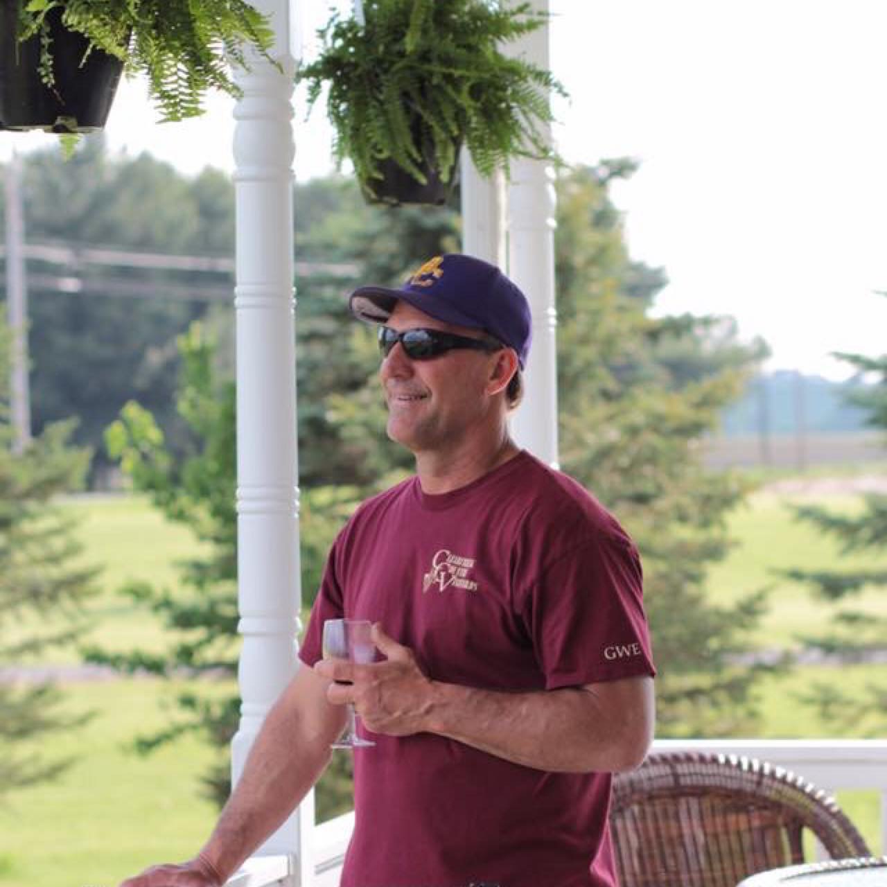 Man wearing sunglasses and a baseball cap holding a glass, standing outdoors near white pillars with greenery in background.