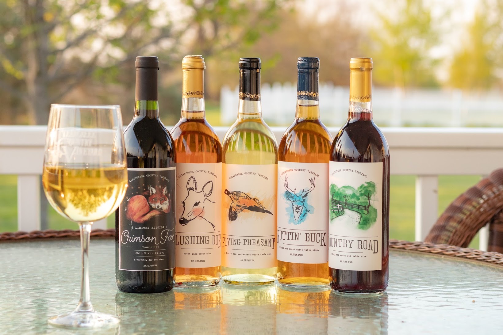 Five bottles of wine and a glass of white wine on a table outdoors with trees in background.