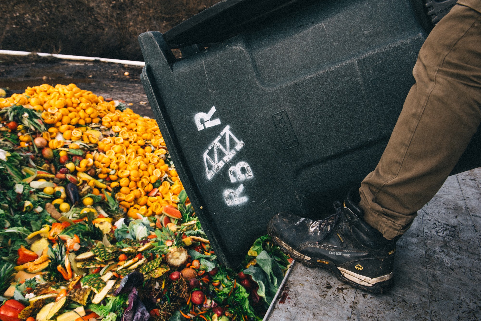 Person pouring produce from a black container onto a large pile of fruits and vegetables outdoors.