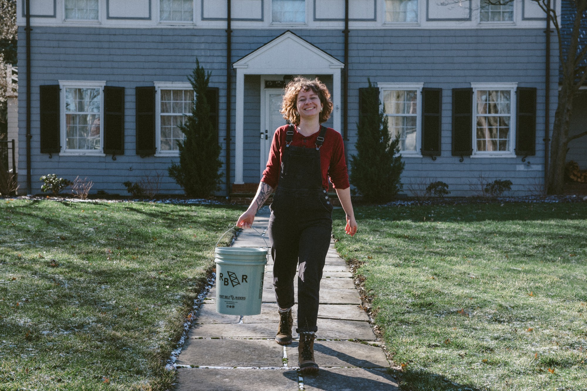 Person with curly hair walking on a sidewalk carrying a bucket, in front of a large house with trees and grass.