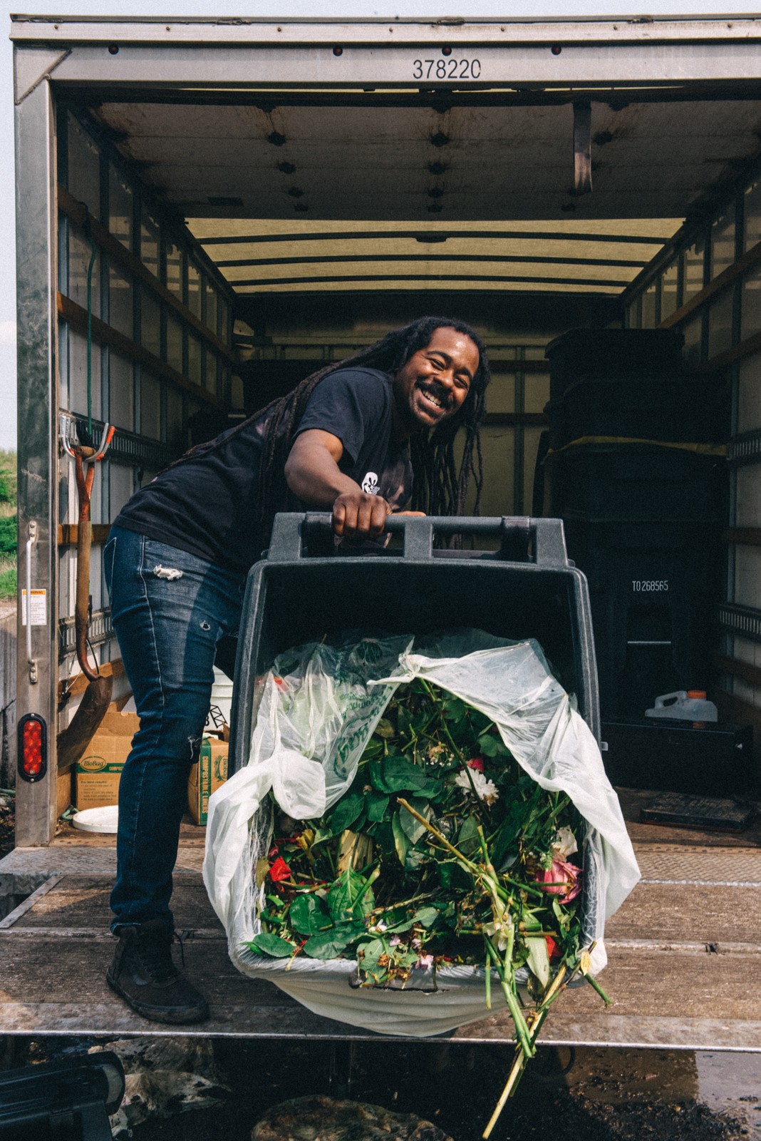 Person with long hair and dark clothing smiling, standing inside a truck with a large container of flowers.