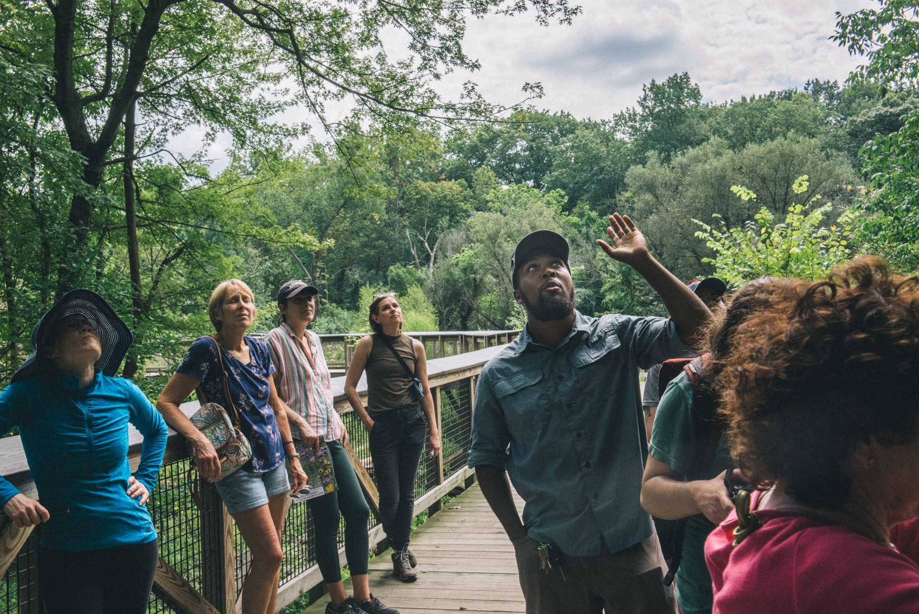Group of people walking on a wooden path surrounded by trees and greenery, some are talking and looking around.
