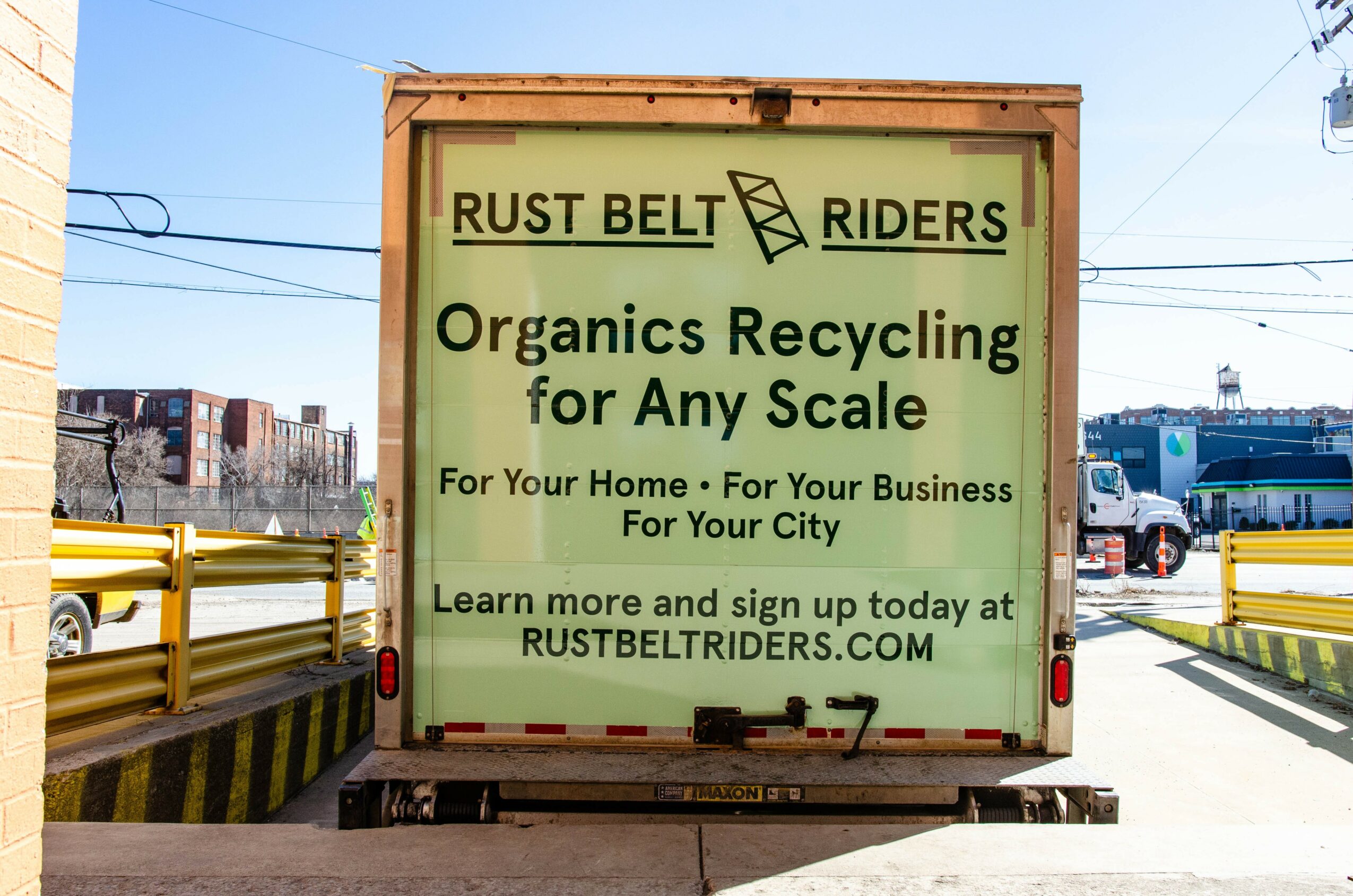 Recycling truck with sign advertising Rust Belt Riders organic recycling service, cityscape background, clear sky.