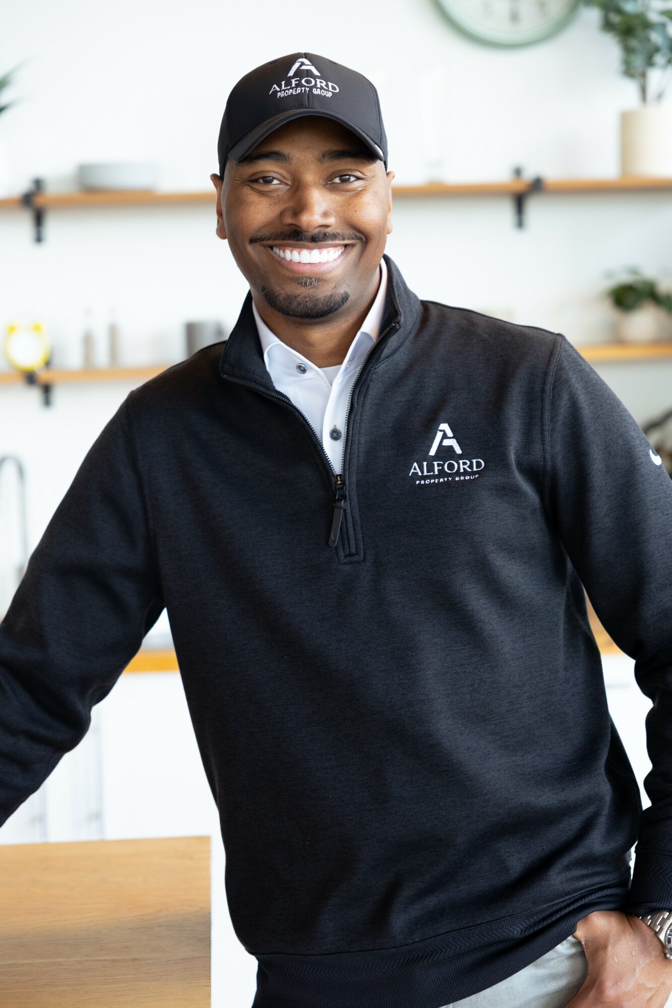 Smiling man in black Alford uniform and cap indoors, shelves and clock in background.