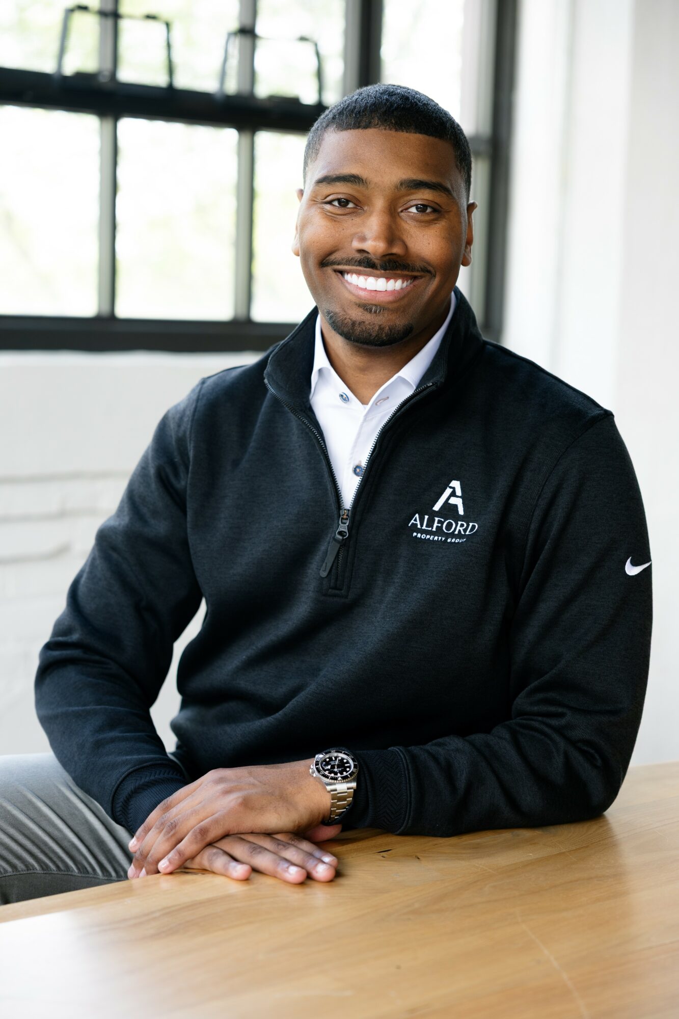 Young man smiling, sitting at a table indoors with large windows in background.
