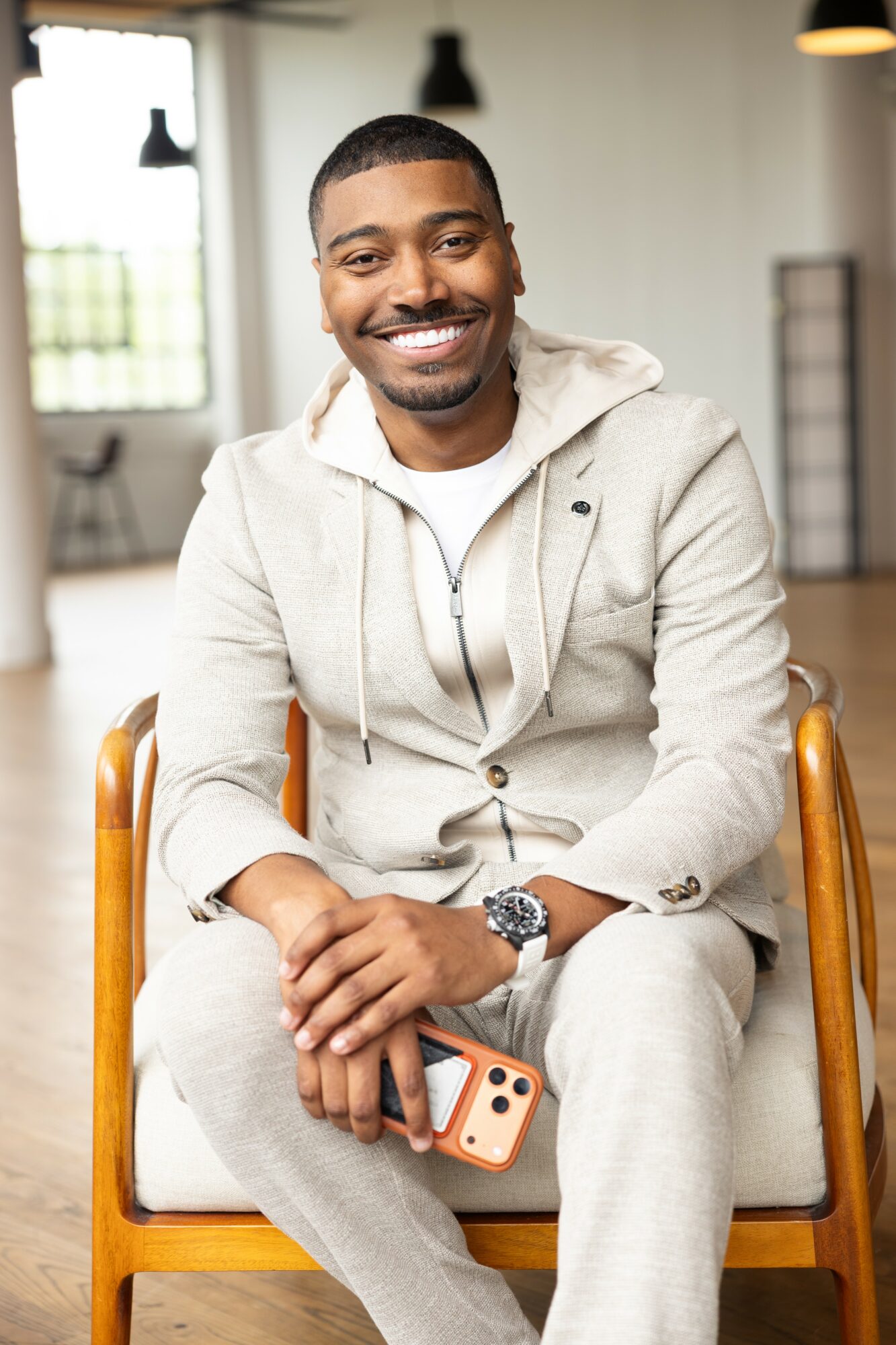 Smiling man sitting on a chair holding a smartphone in a bright room with large windows.