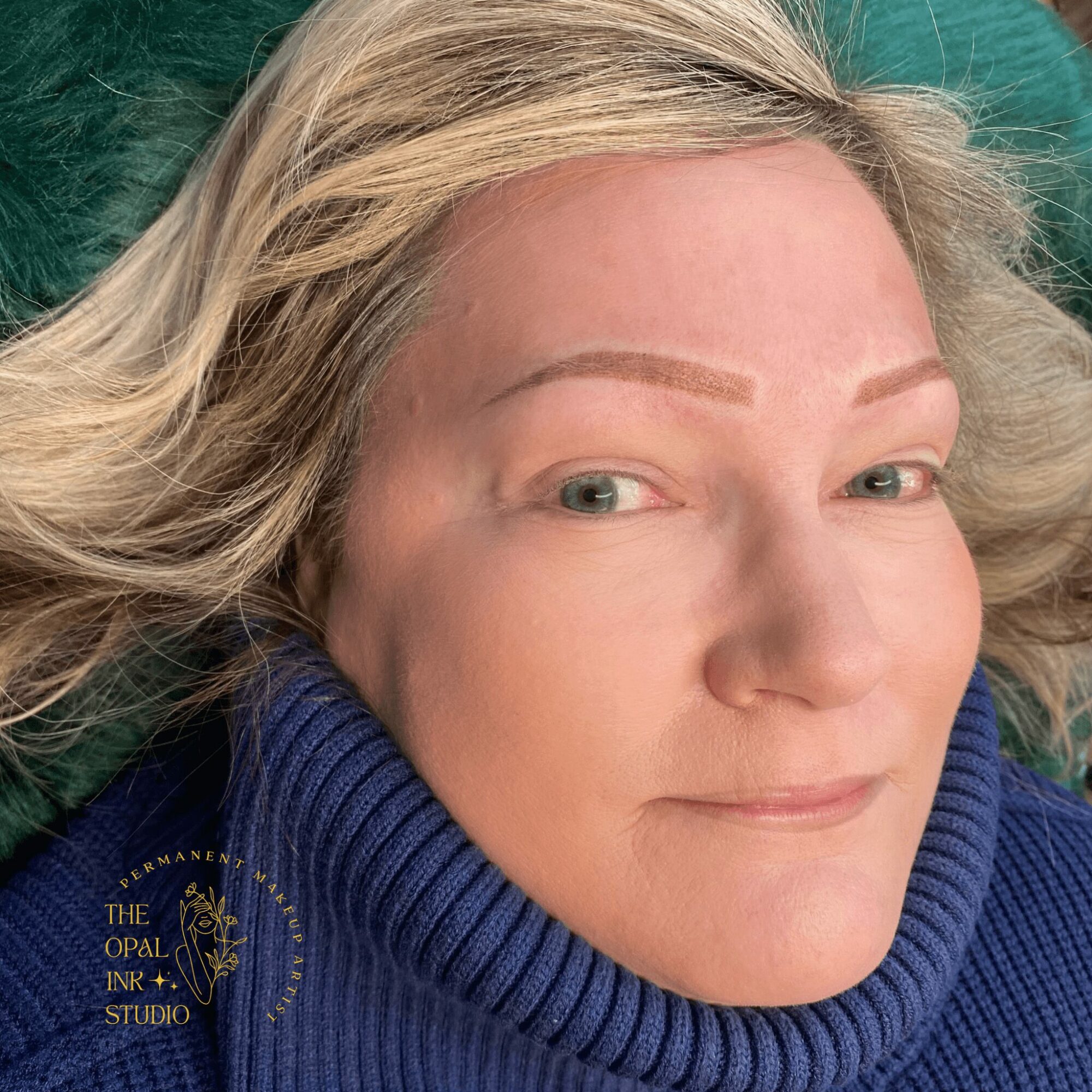 Close-up of a woman with blonde hair lying on a green surface, wearing a blue turtleneck sweater.