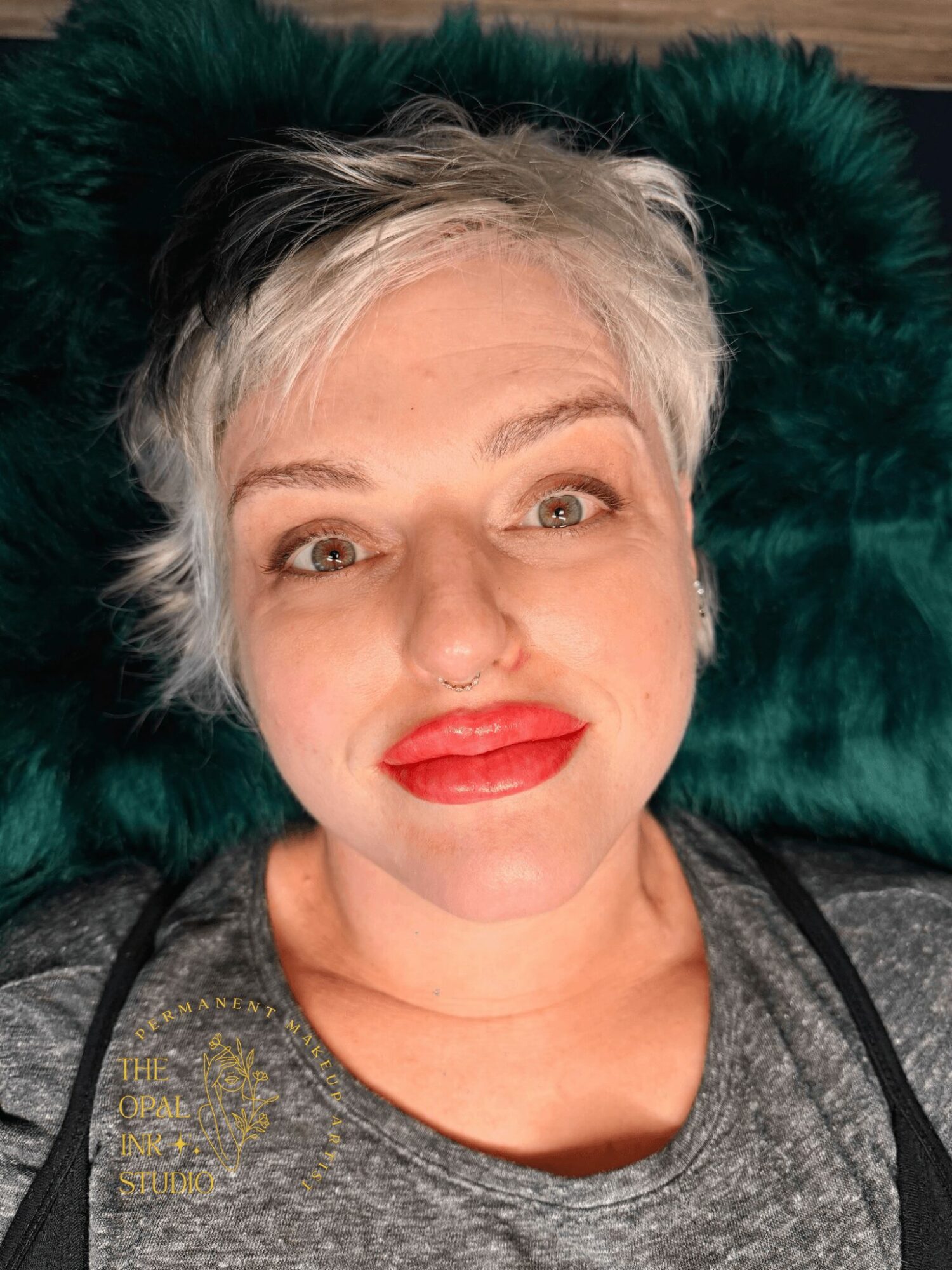 Woman with short gray hair lying on a dark green furry surface, smiling with red lipstick, wearing a gray shirt.