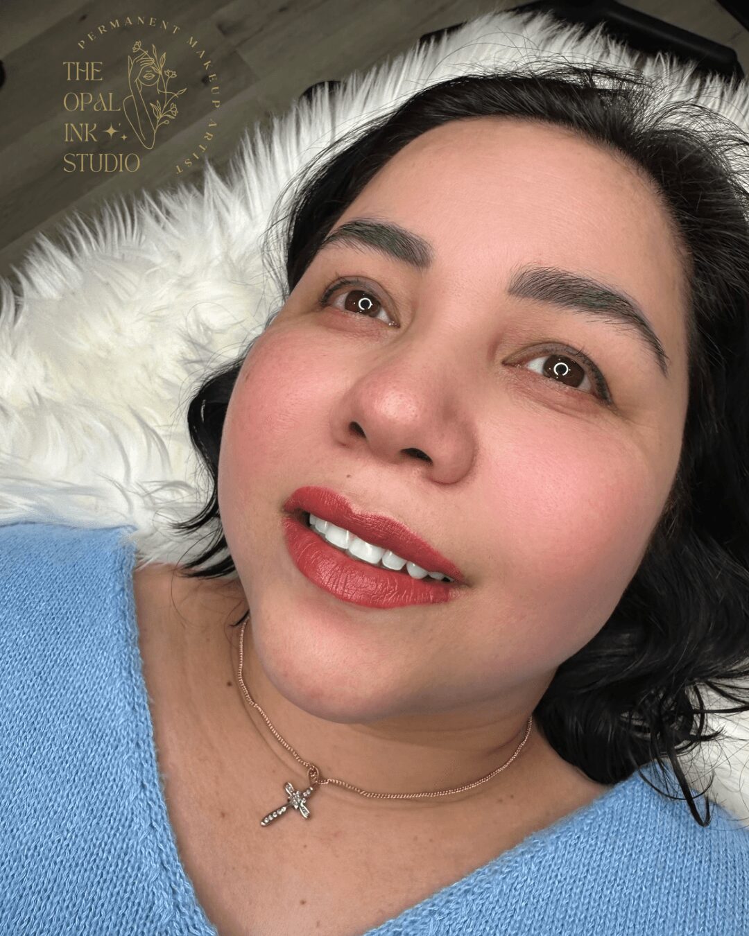 Close-up of a woman lying on a white furry surface, smiling, with dark hair, wearing a blue top and a cross necklace.