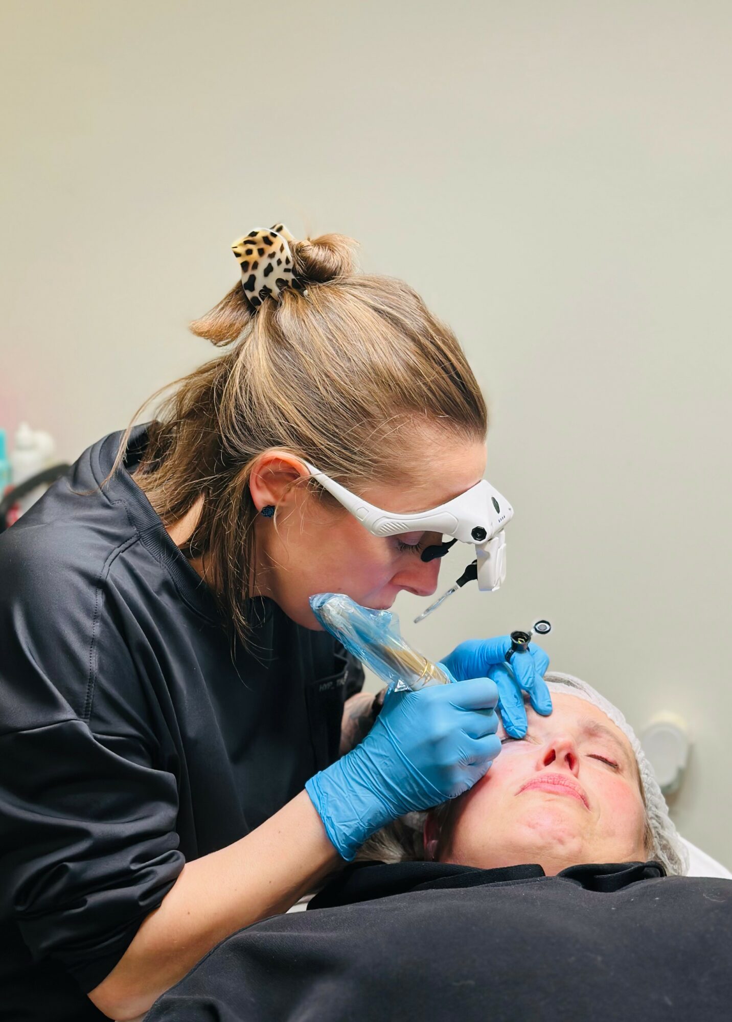A woman in protective glasses and blue gloves performs a cosmetic procedure on a person's face in a clinical setting.