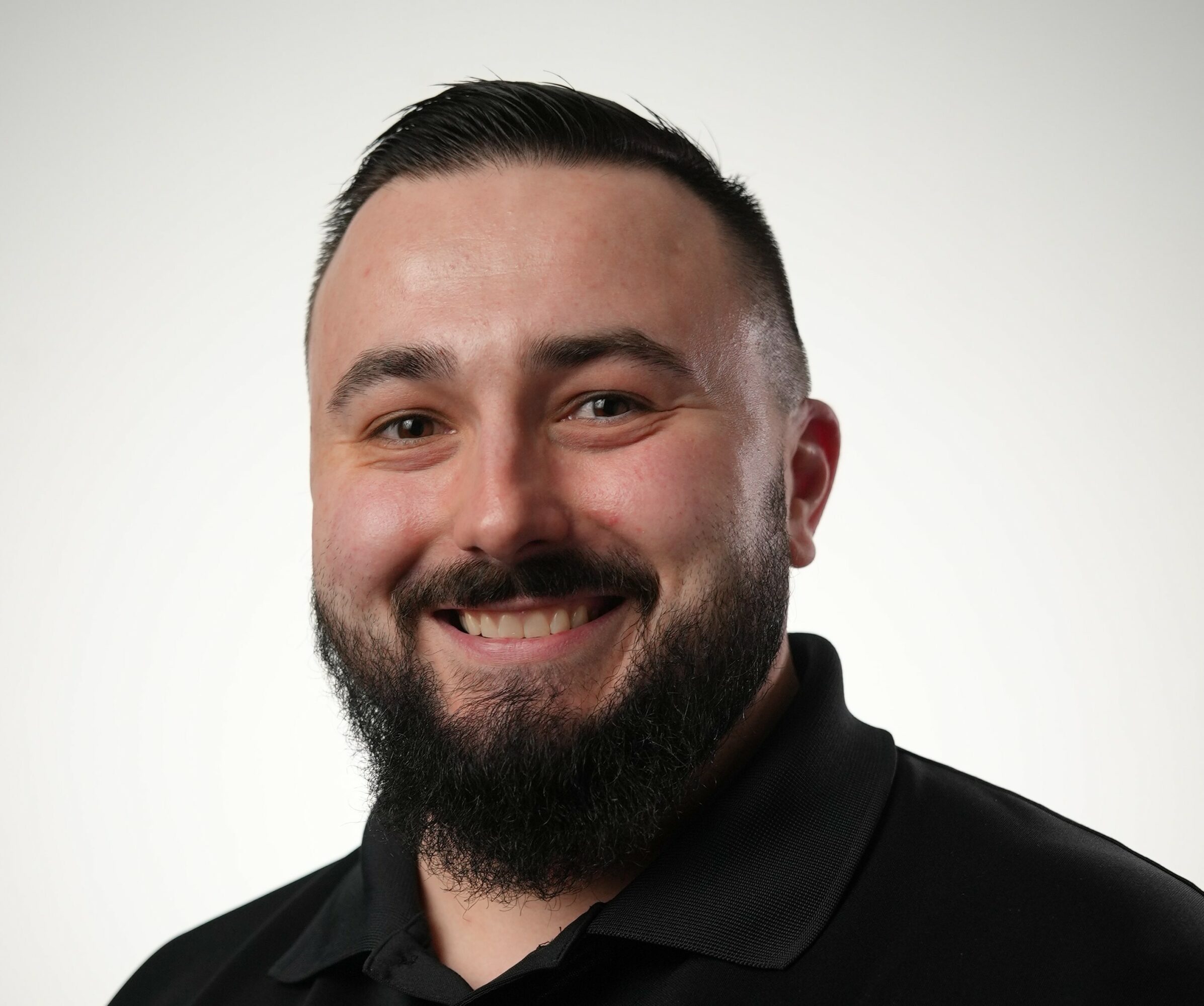 Smiling man with dark hair and beard, wearing a black shirt, against a plain light background.