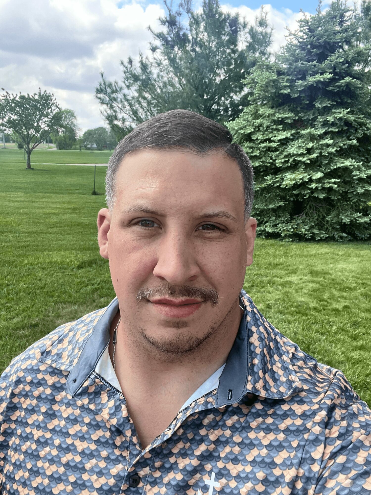 Young man with short dark hair and a mustache outdoors, wearing a patterned shirt, with trees and grass in the background.