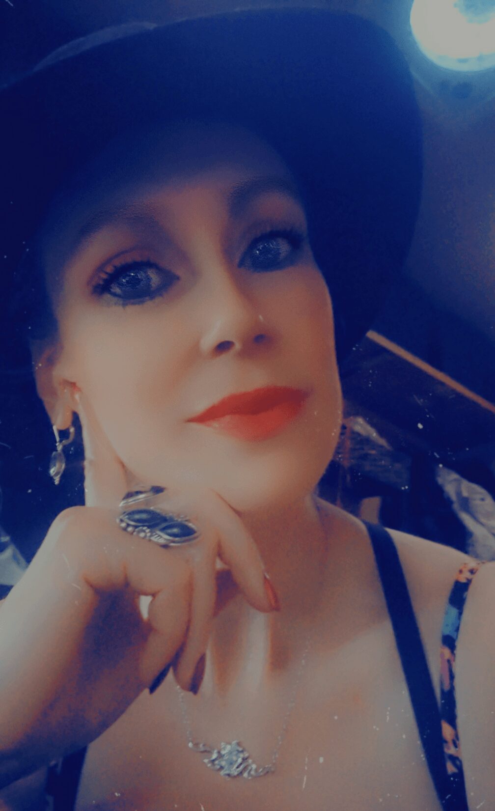 Woman with makeup, wearing a hat and earrings, resting her chin on her hand, looking at the camera.