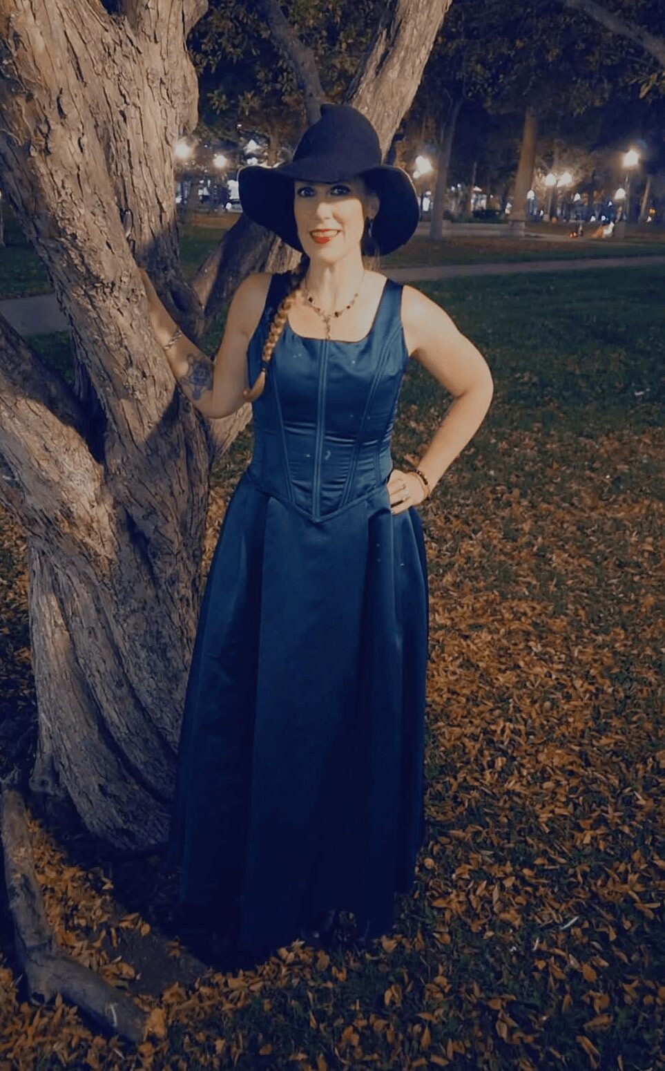 Woman in a long blue dress and wide-brimmed hat standing outdoors near a tree at night.