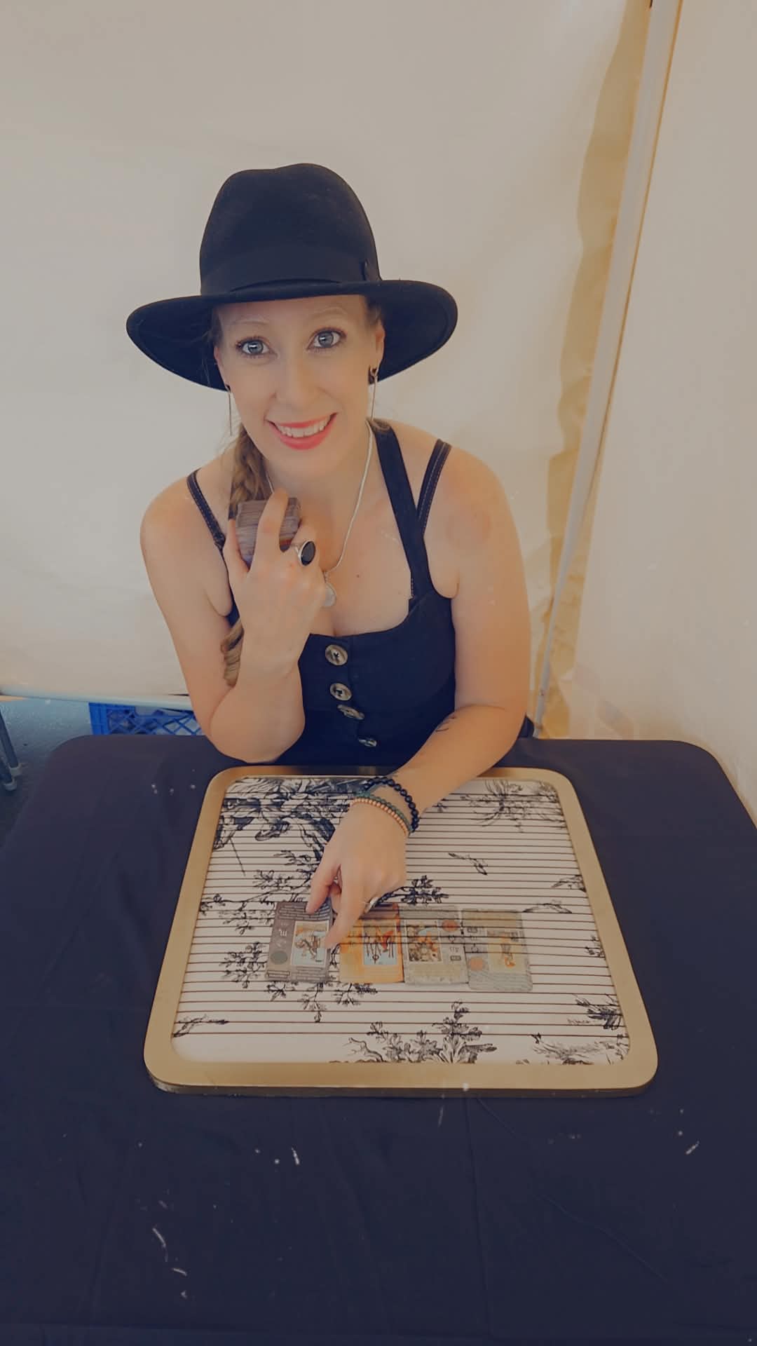 Woman wearing a black hat and sleeveless top, sitting at a table with a musical instrument, smiling at the camera.