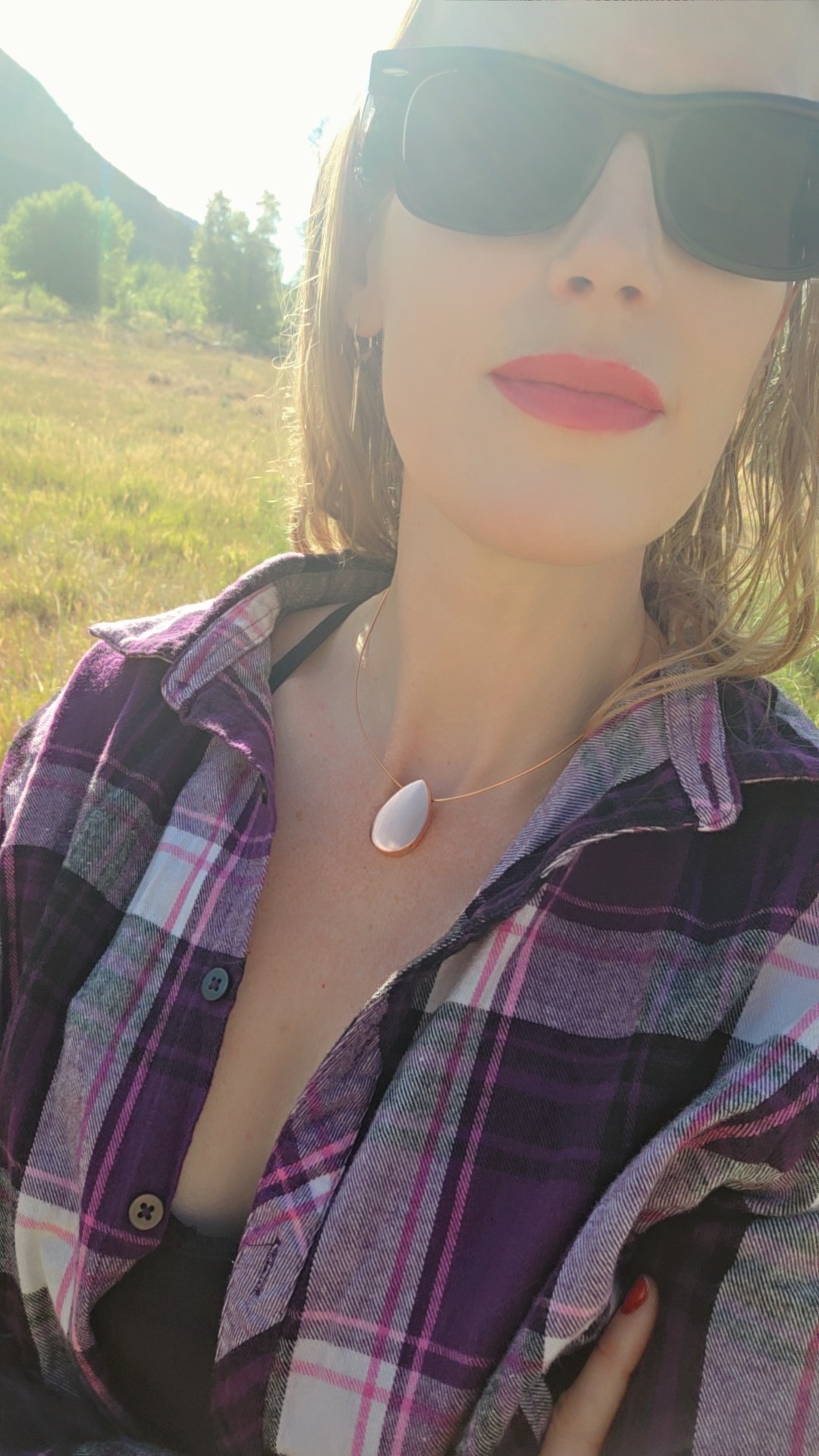Woman wearing sunglasses and a plaid shirt outdoors with mountains and greenery in background.