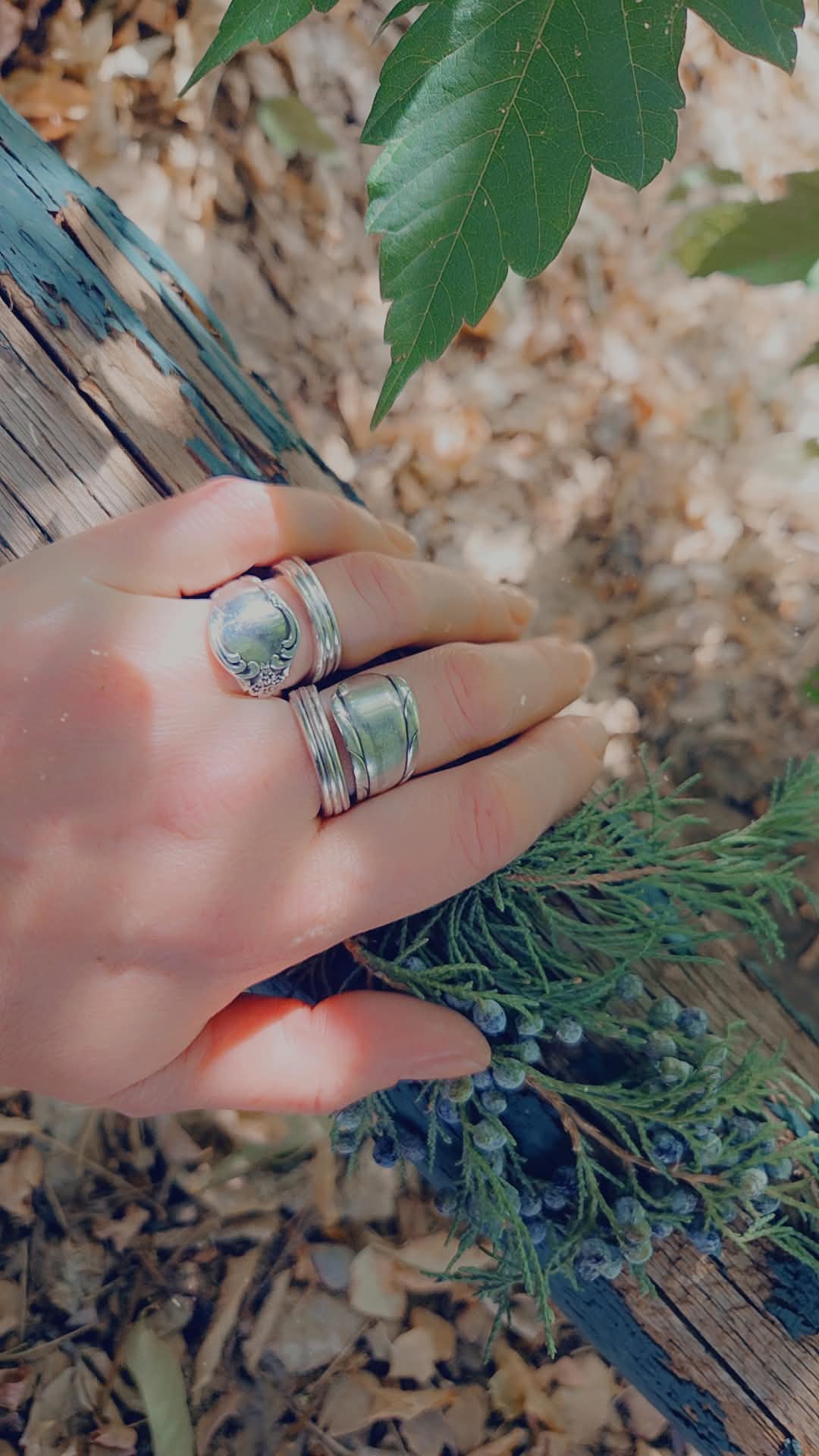 Hand with multiple rings resting on a wooden surface surrounded by green leaves and plants.