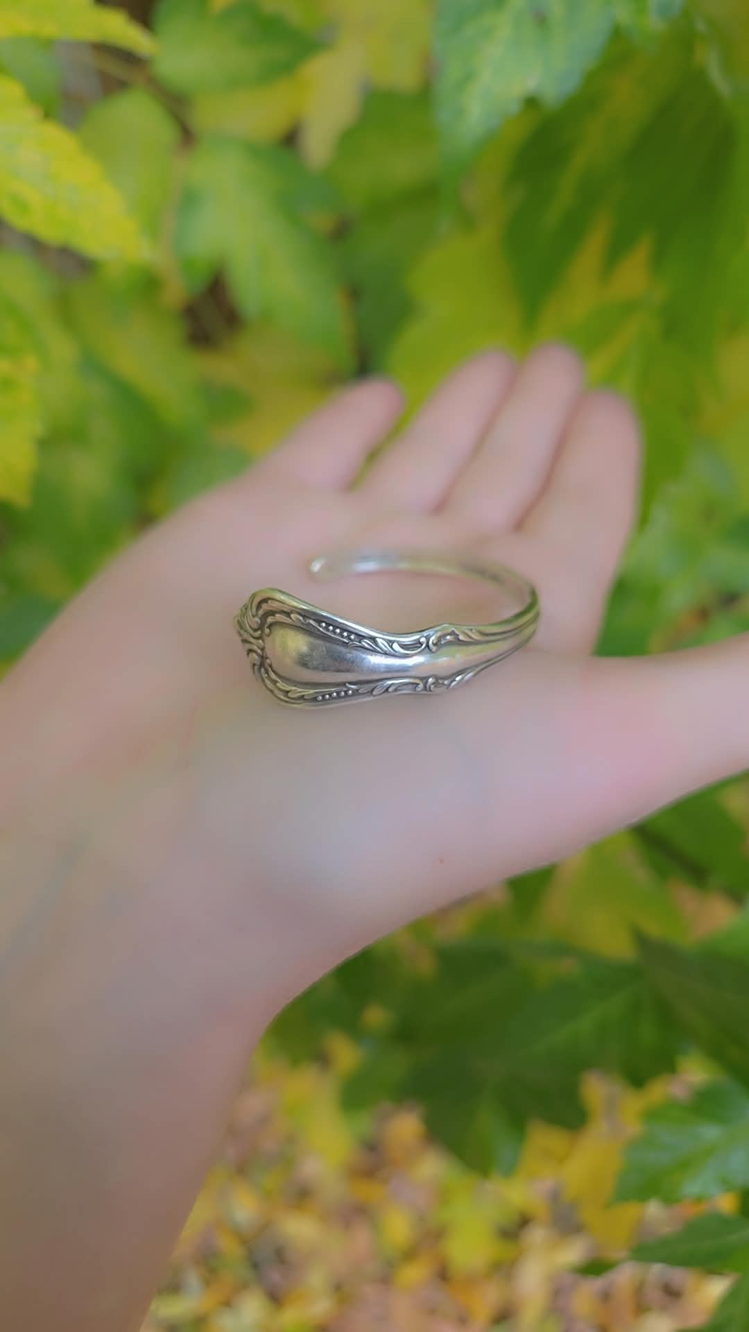 Person's hand holding a silver ring with detailed design, green and yellow leaves in background.