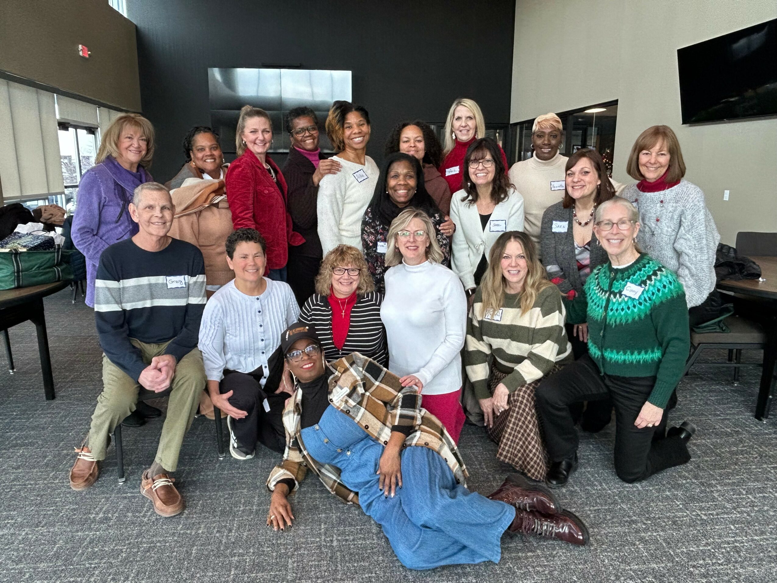Group of 16 diverse people posing indoors, some sitting and some standing, smiling at the camera.